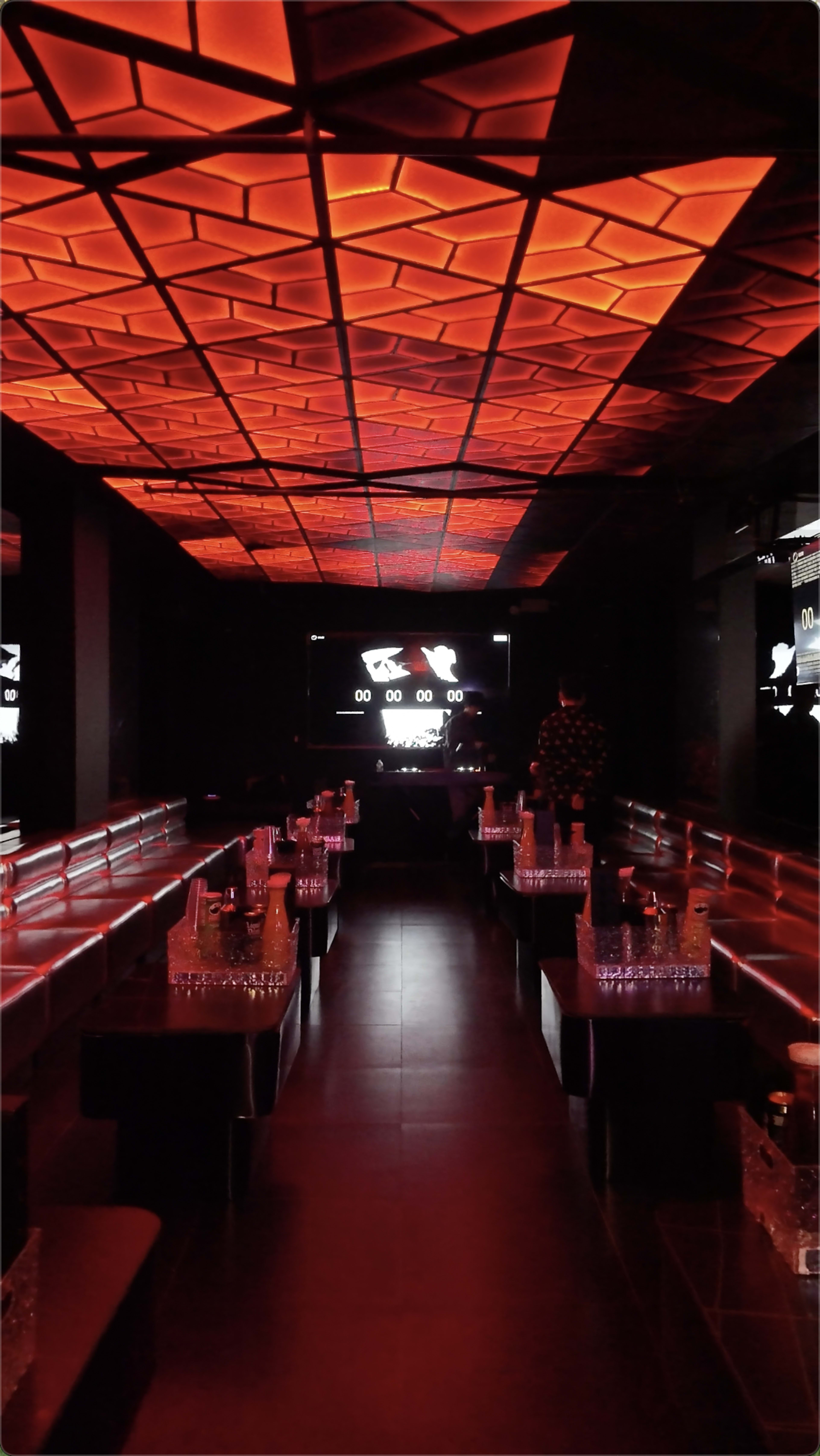 The image depicts a dimly lit nightclub with red lighting, featuring long benches and a large screen displaying a countdown clock.