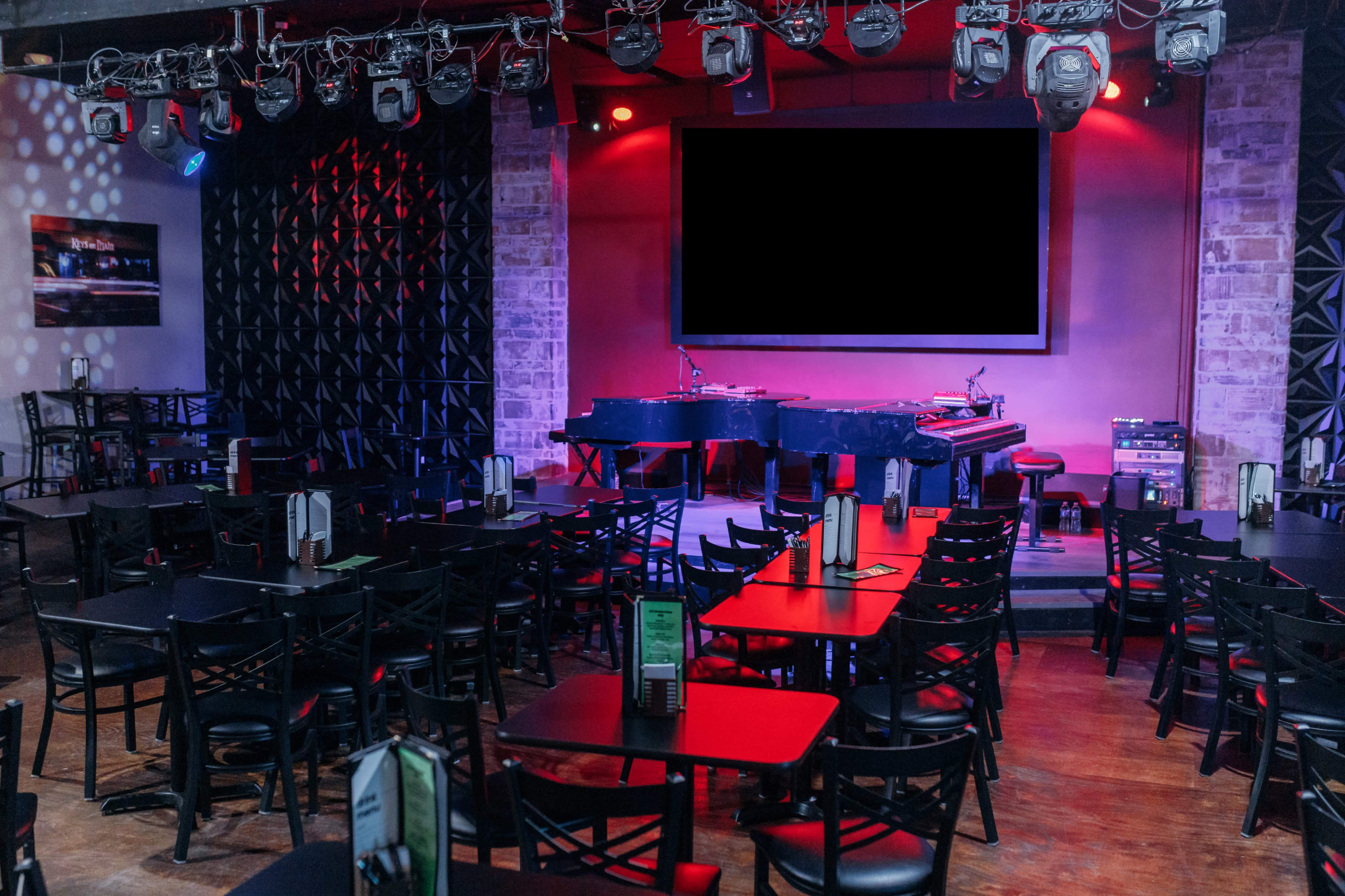 An empty performance venue with rows of black tables and chairs facing a small stage that features a grand piano and a large screen.