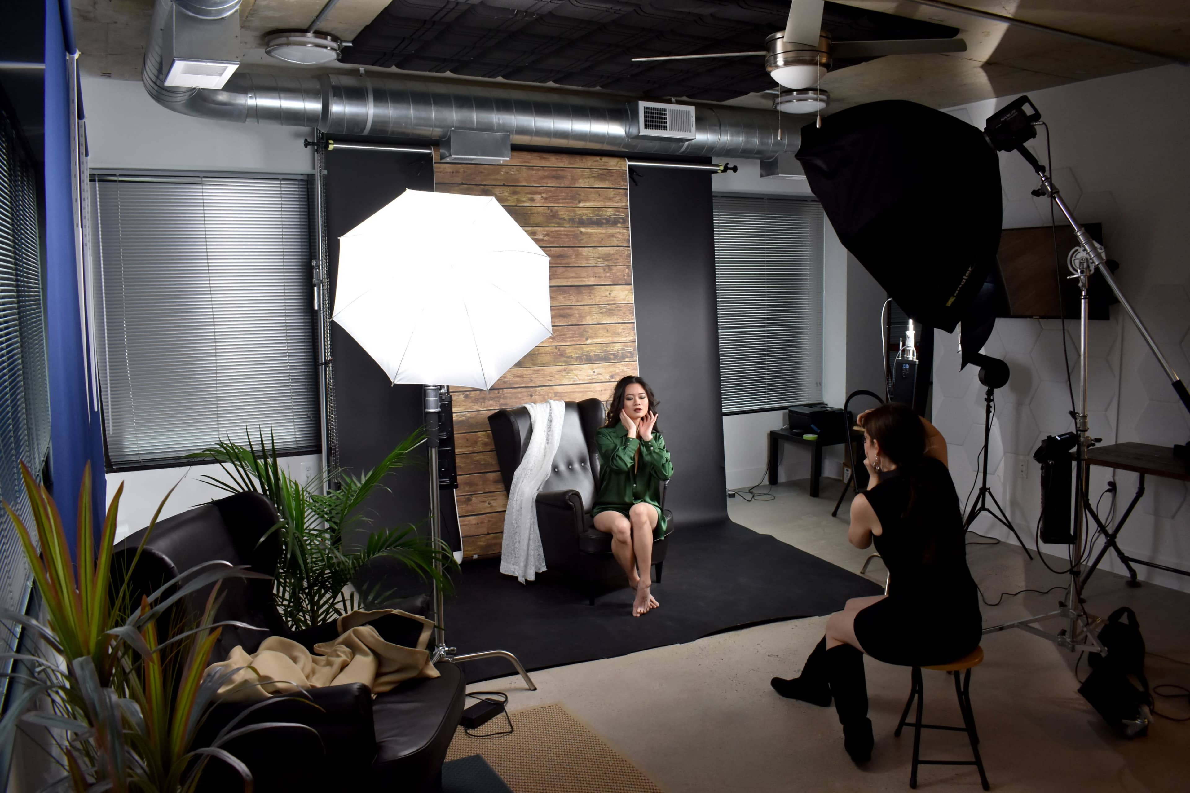 A model sits on a chair in a photography studio, surrounded by soft lighting equipment and backdrops, while another person adjusts the scene.