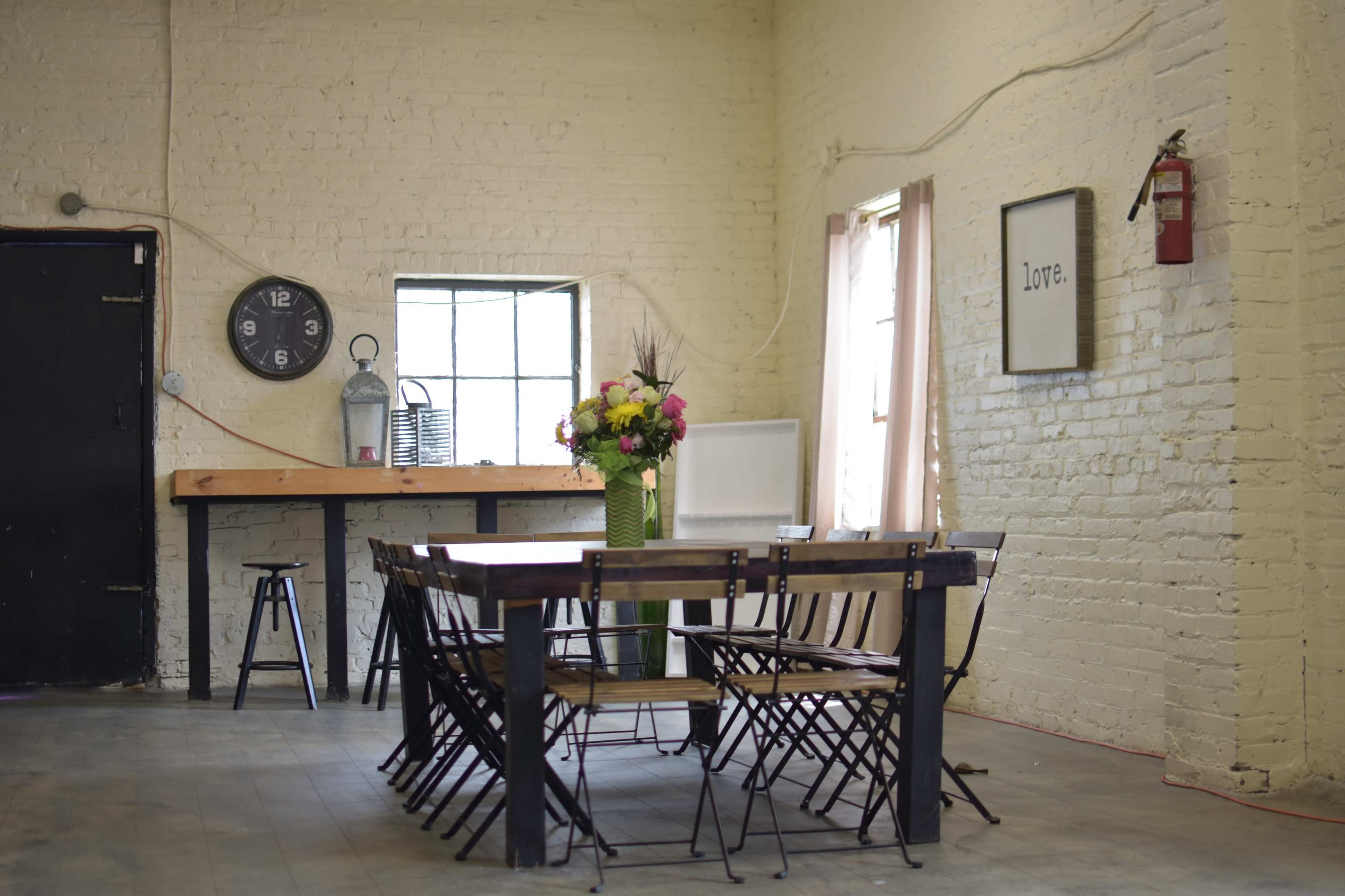 A large, simple dining table surrounded by folding chairs is set in a bright, minimalist room with brick walls and a clock on the wall.