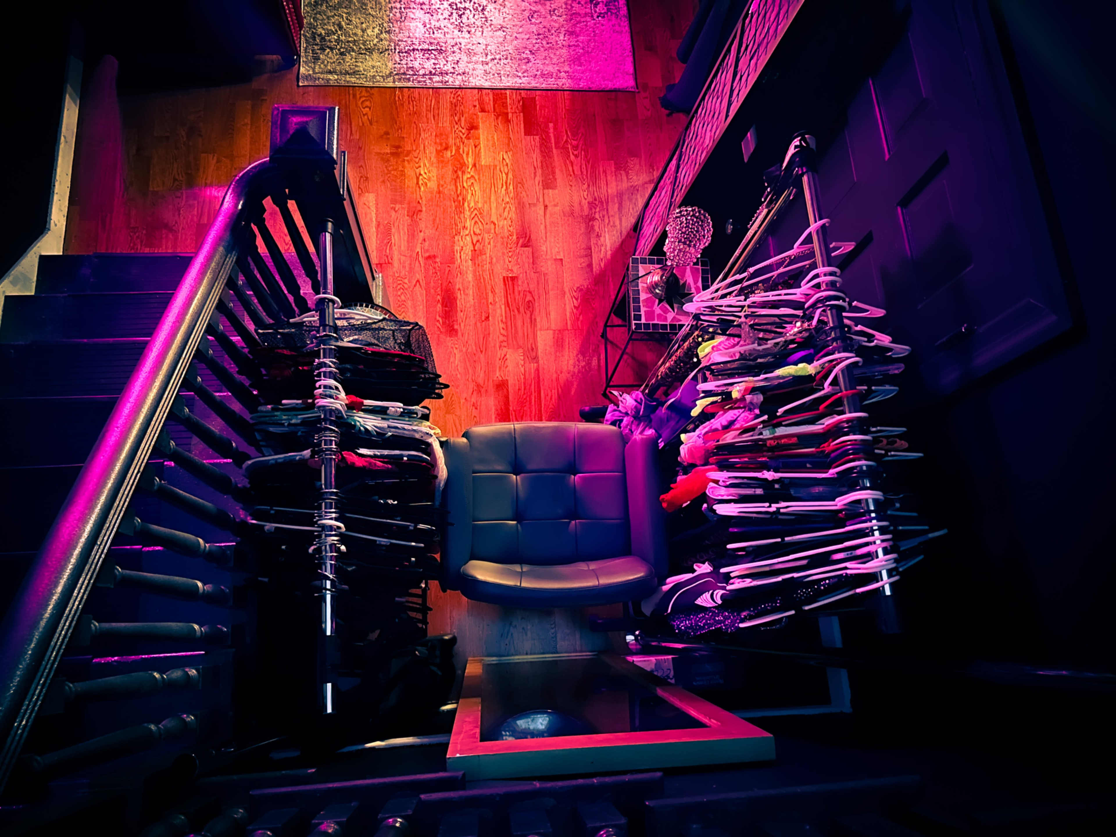 A stairway with a chair at the bottom, flanked by racks of clothing on either side.