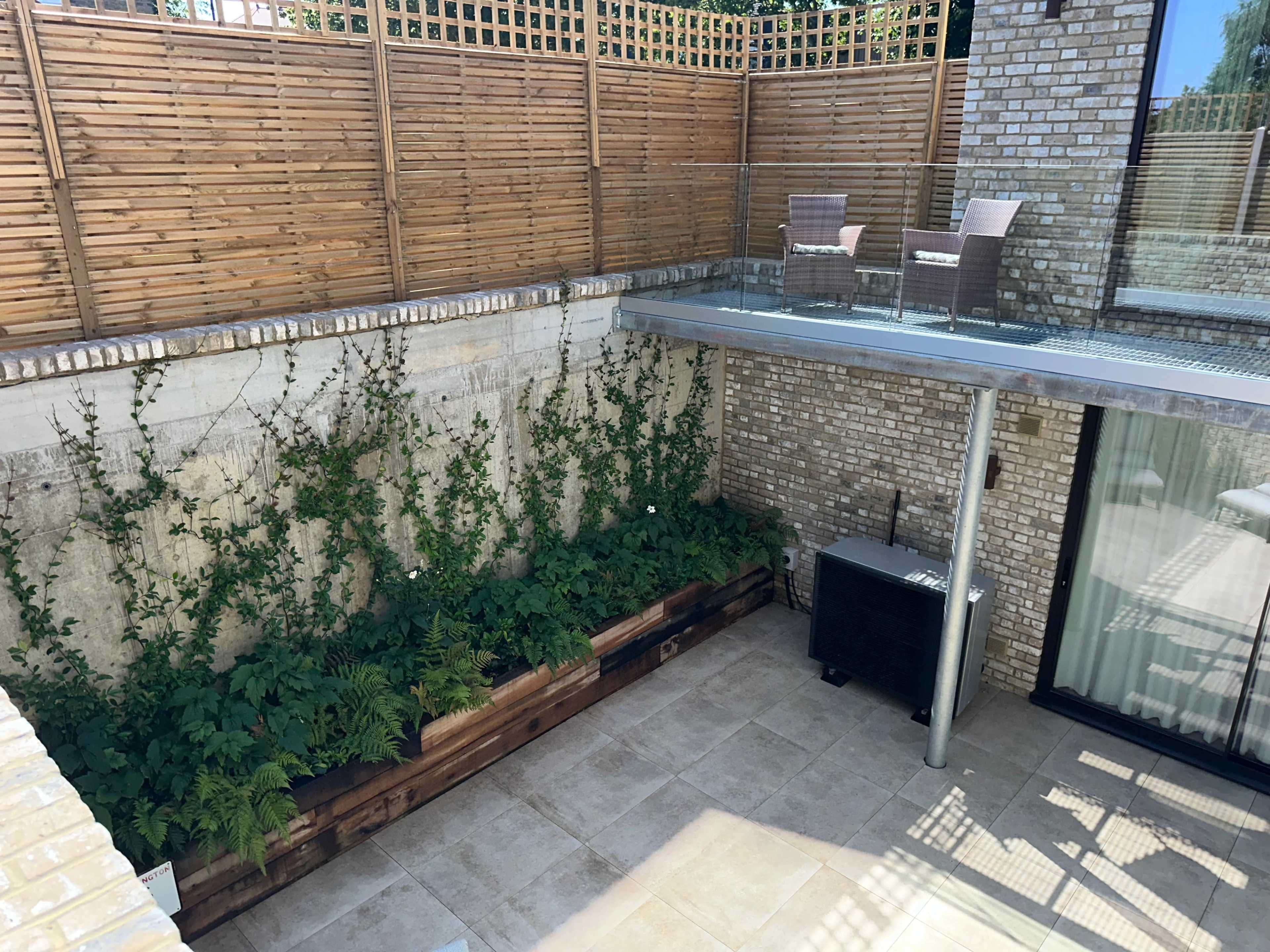 The image shows a modern outdoor space with wooden fencing, a glass railing, and a corner seating area, surrounded by lush green plants in a raised planter.