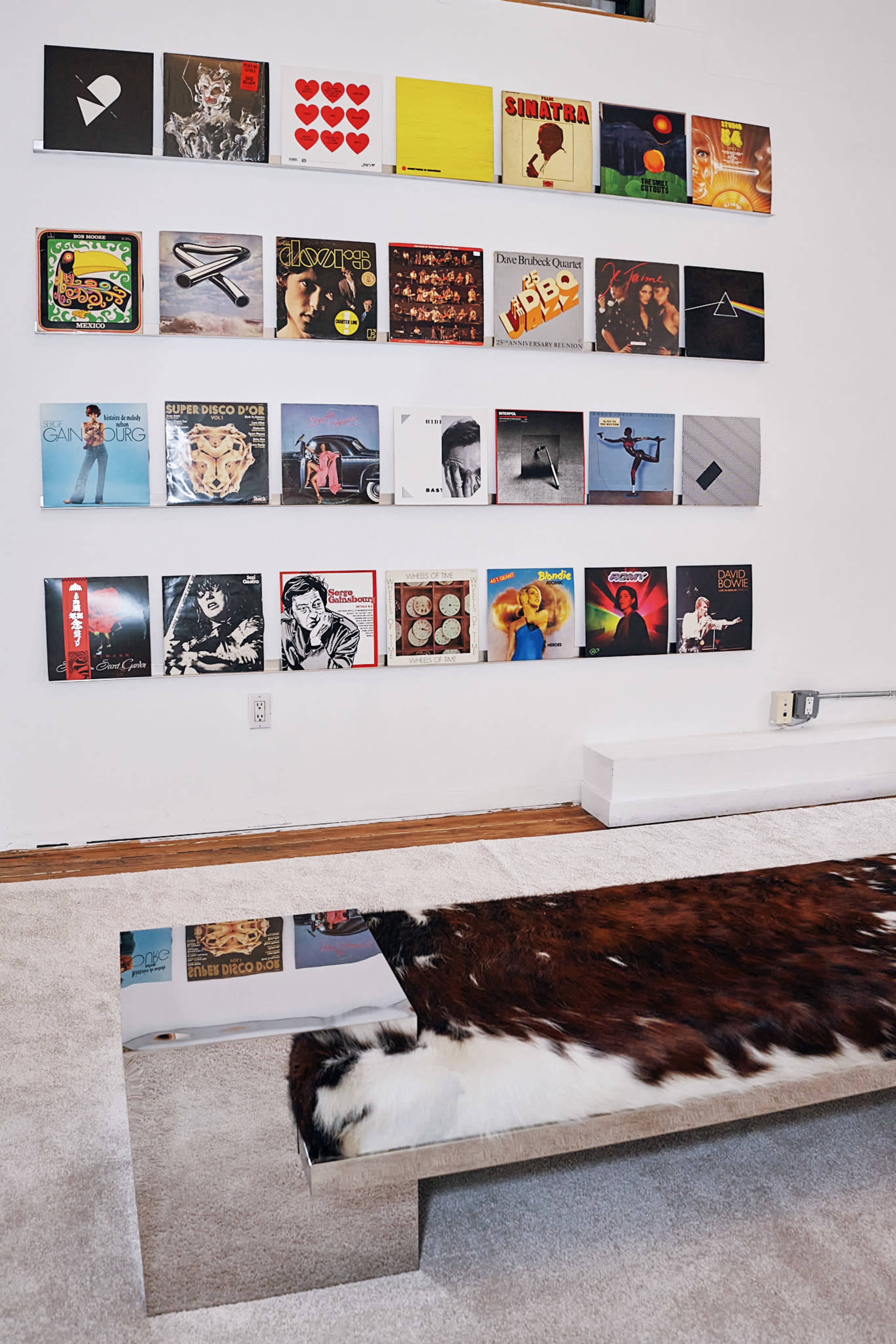 The image features a wall display of various vinyl records arranged in a grid pattern above a low table with a mirrored surface and a textured brown and white rug.