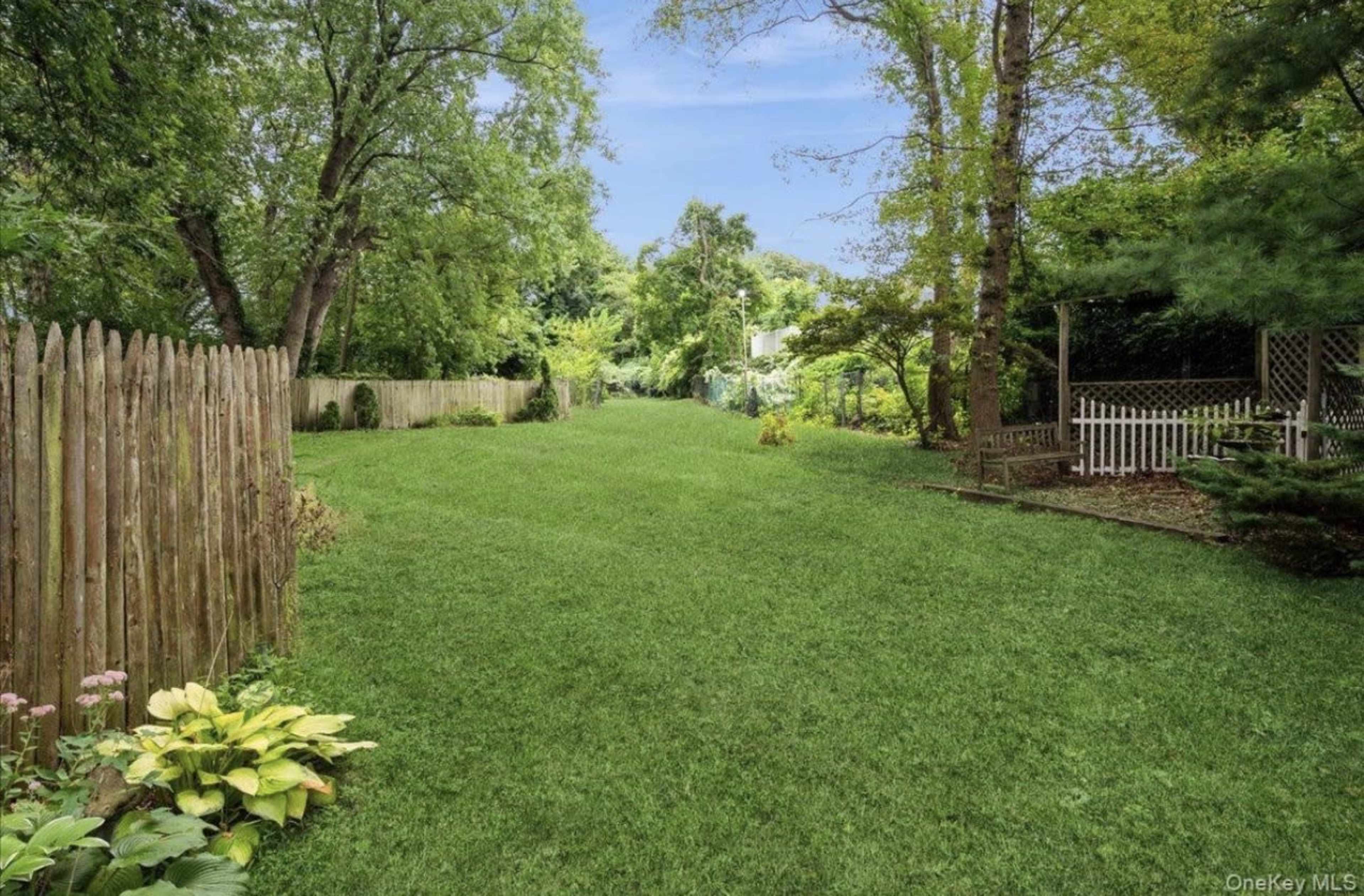 A wide, grassy backyard is bordered by a wooden fence and features trees and garden elements along the sides.