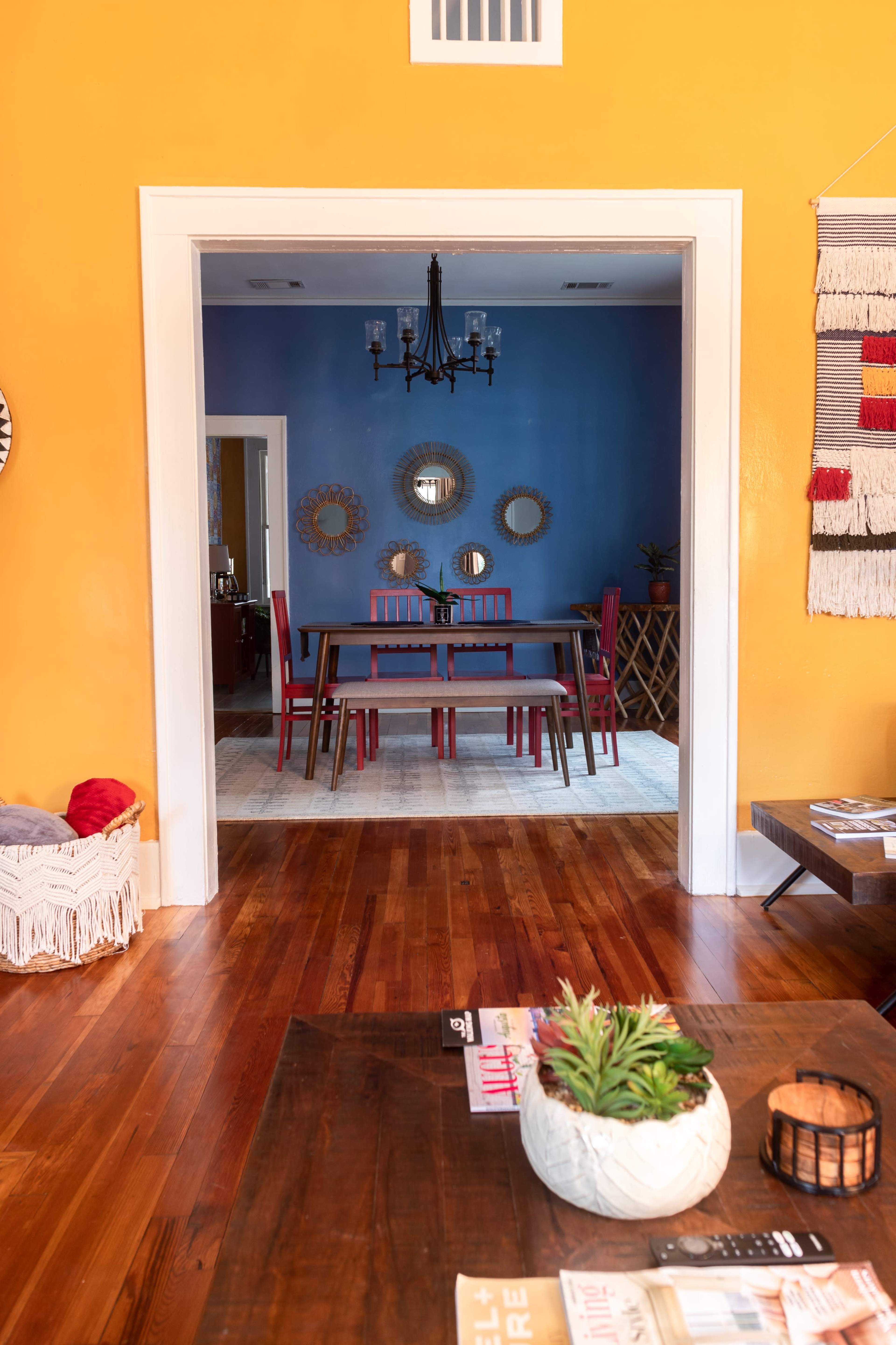A bright yellow wall frames a view of a dining area with a blue wall, featuring a table surrounded by red chairs and decorative mirrors.