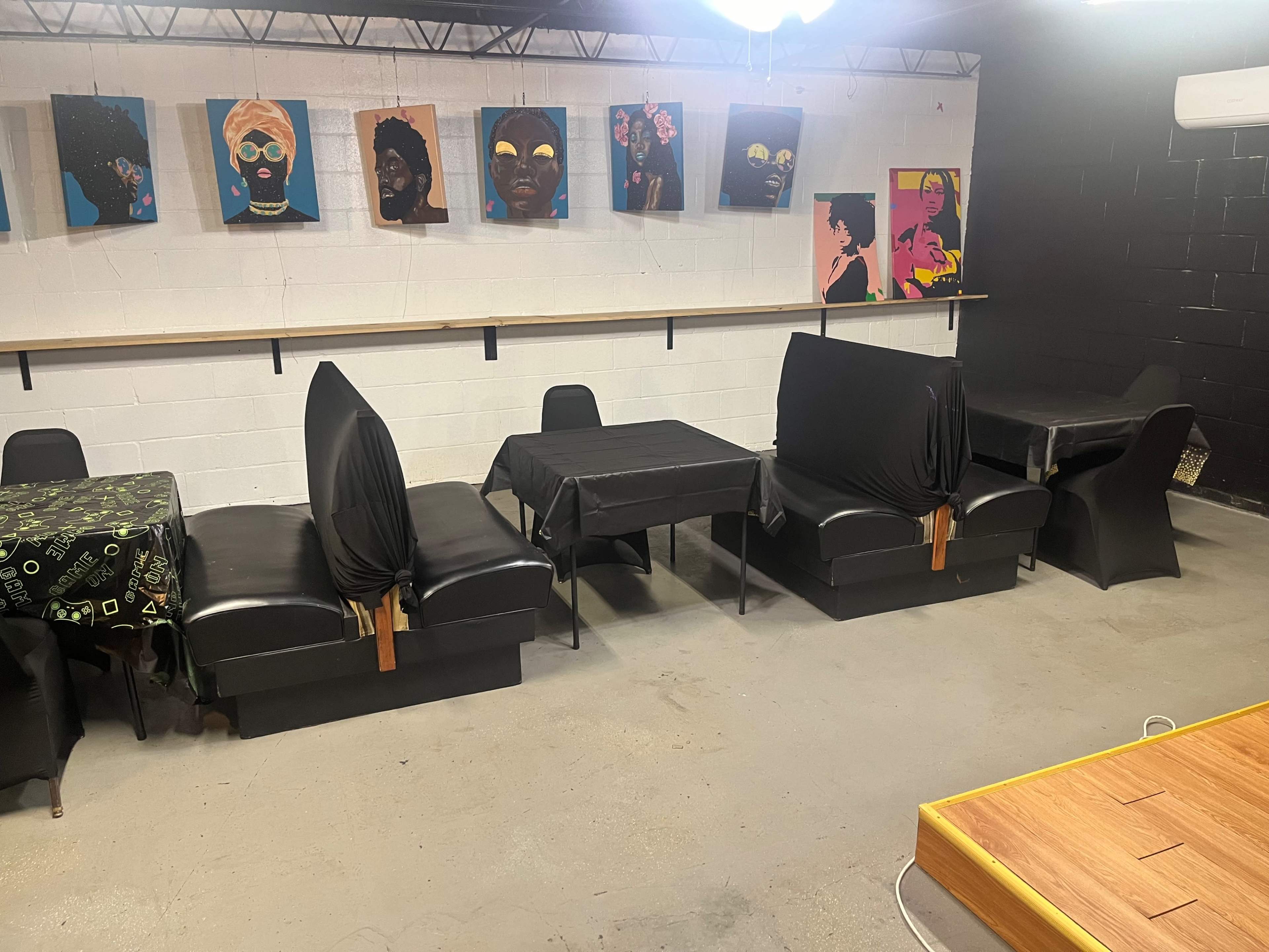 The image shows a room with several black-covered tables and chairs arranged along a wall with colorful artwork.