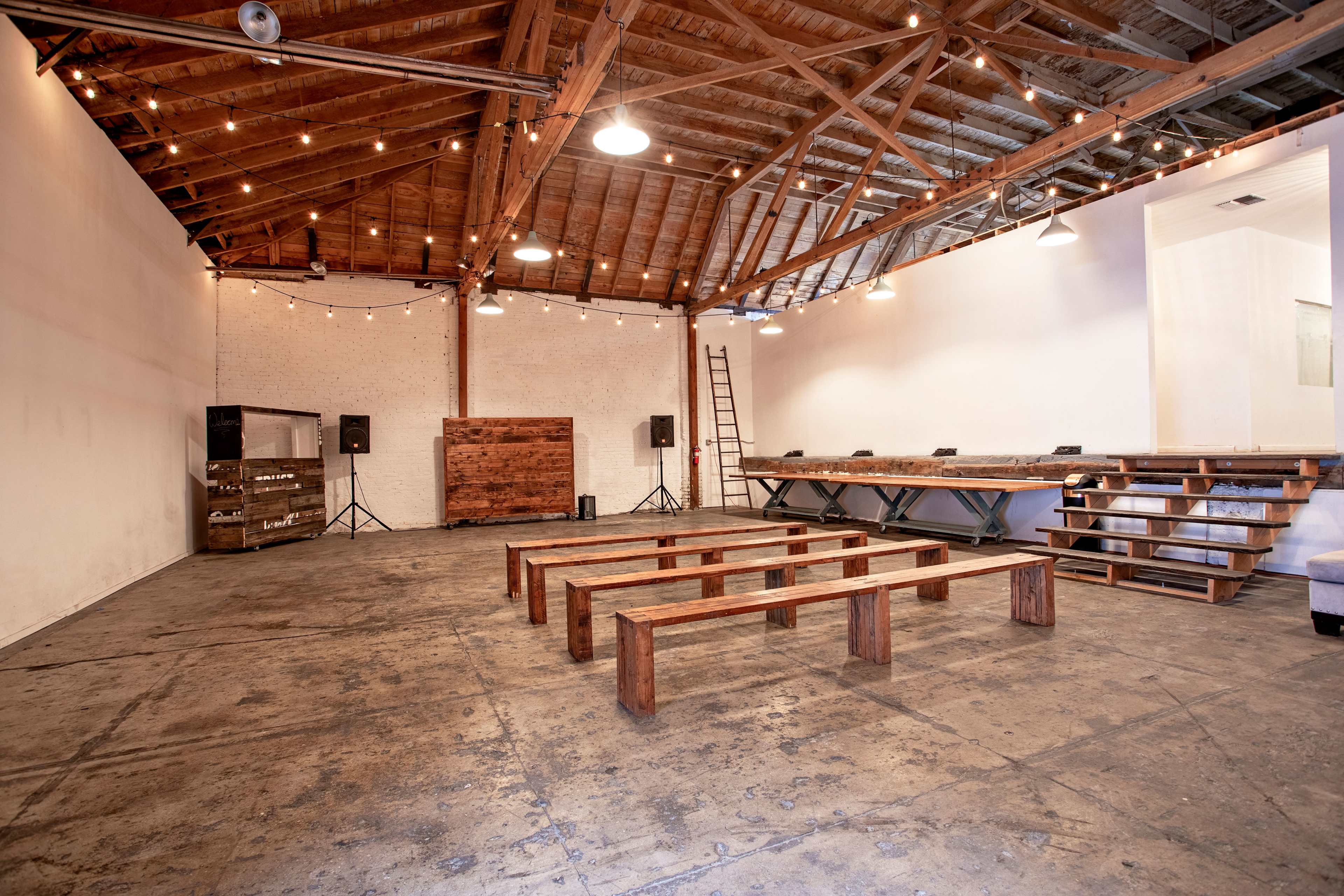 A spacious venue with wooden benches arranged in rows, a raised platform at one end, and string lights hanging from the ceiling.