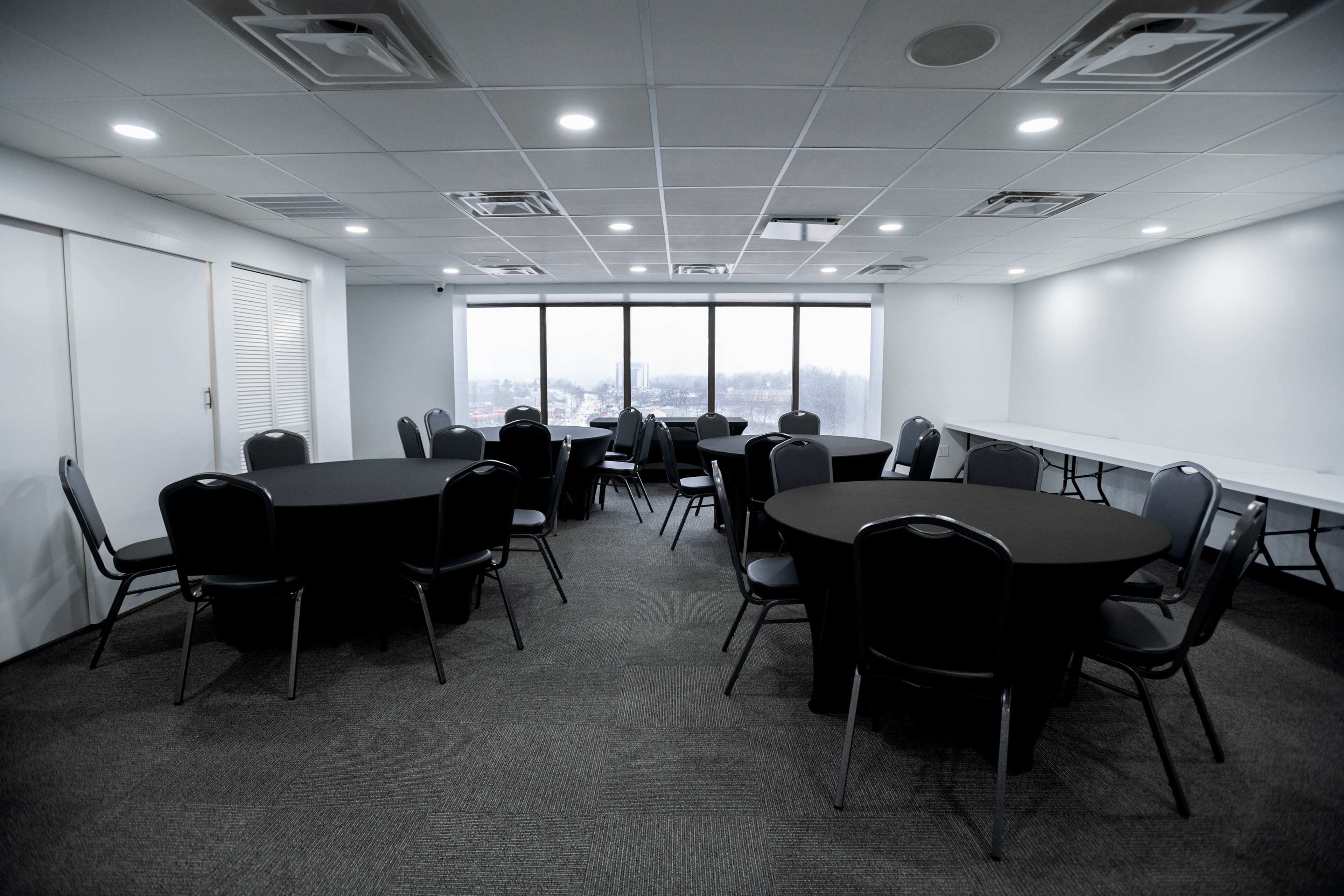 A spacious room with several round tables covered in black linens and a row of windows allowing natural light.