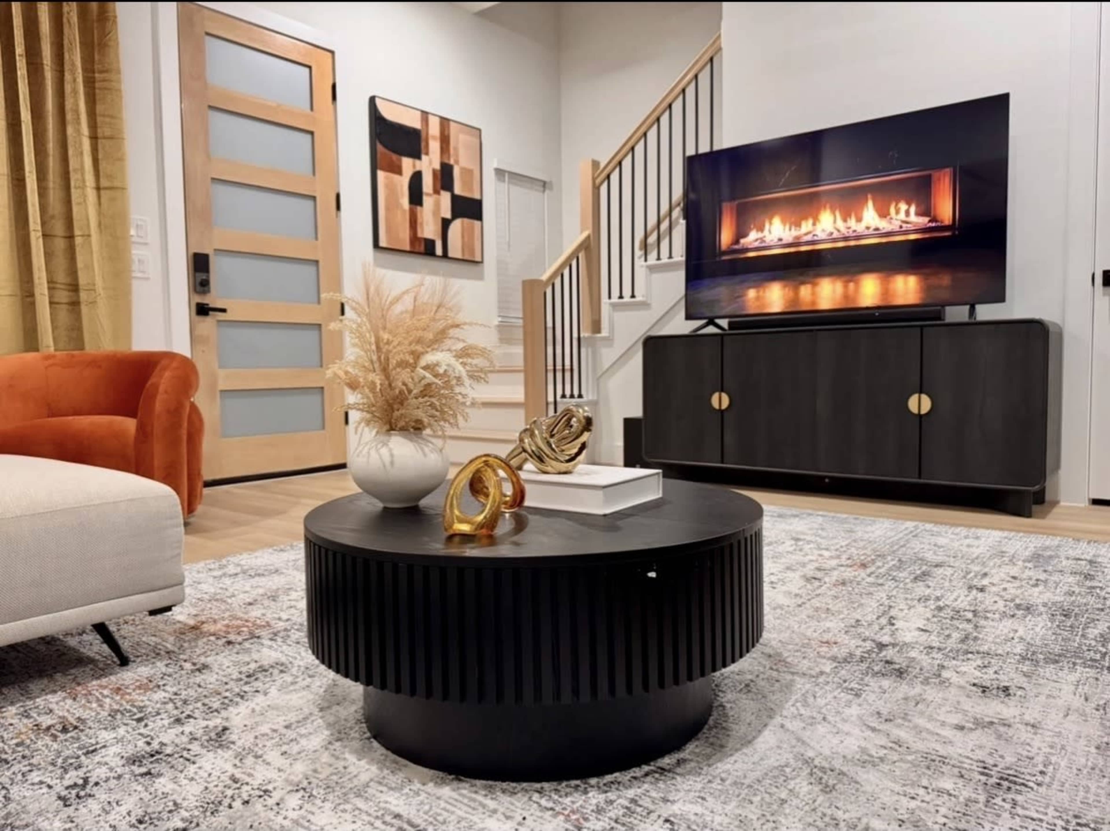 The image shows a modern living room featuring a round black coffee table, a cozy orange armchair, a fireplace on a television screen, and a decorative wall art piece.