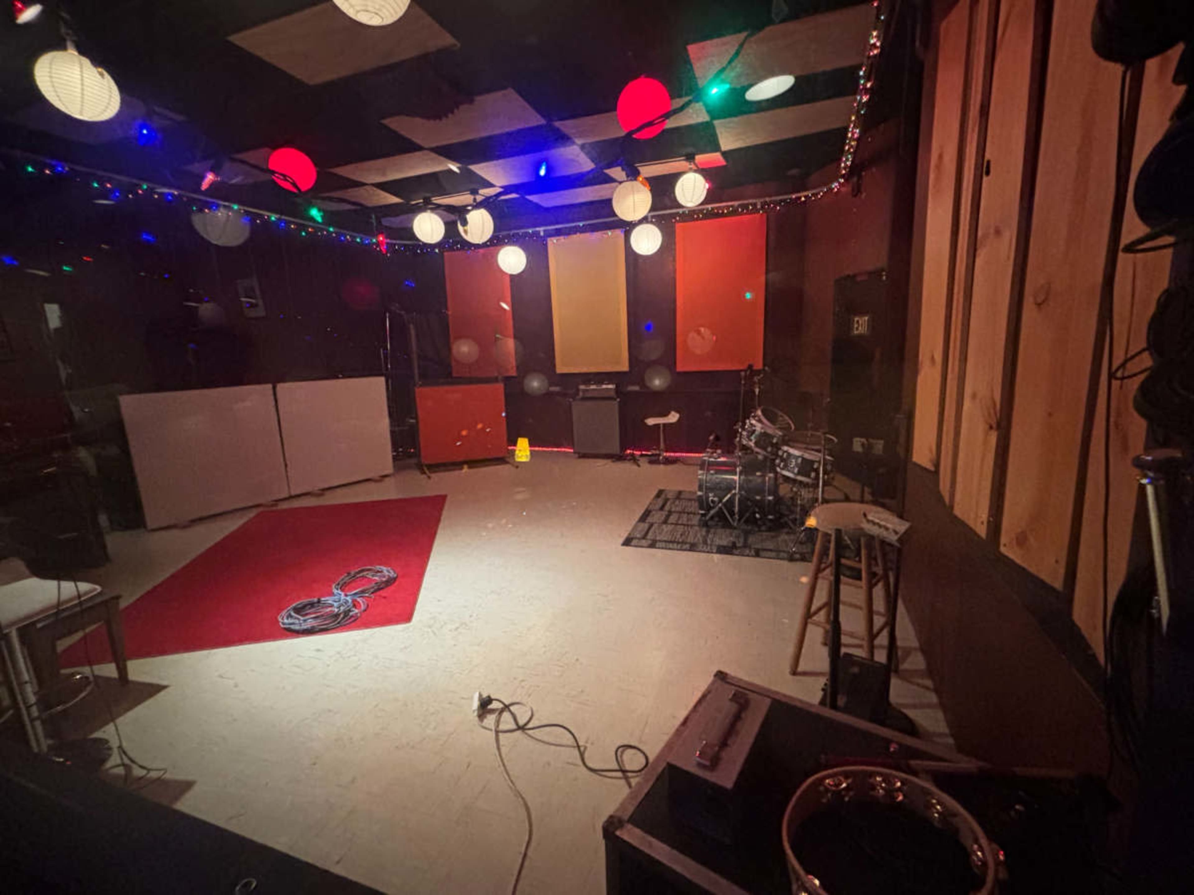 The image shows a music rehearsal space with colorful hanging lanterns, a red area rug, and a drum set positioned on a light-colored floor.