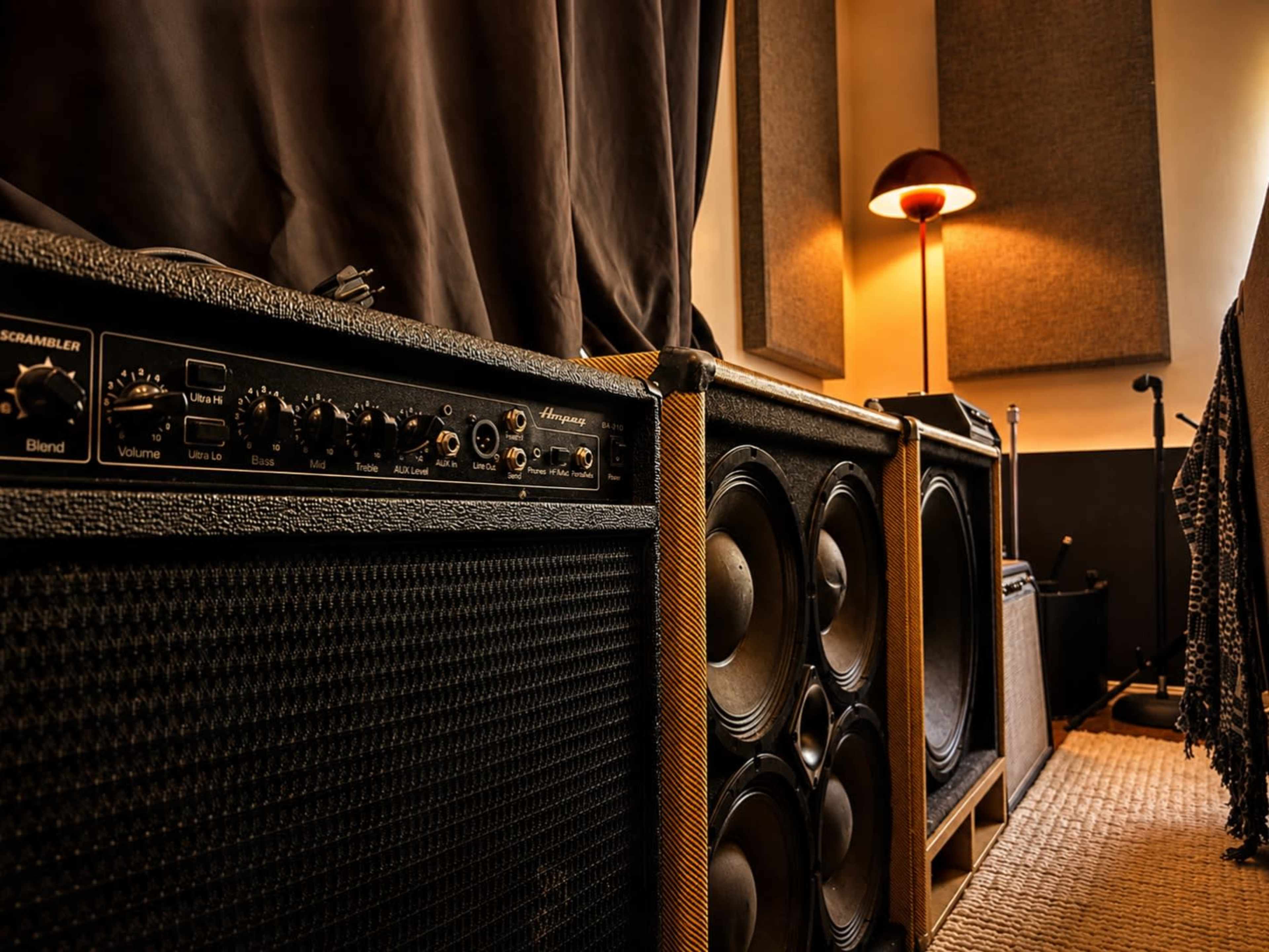 A row of black and wooden amplifiers and speakers is positioned in a dimly lit room with a floor lamp in the background.