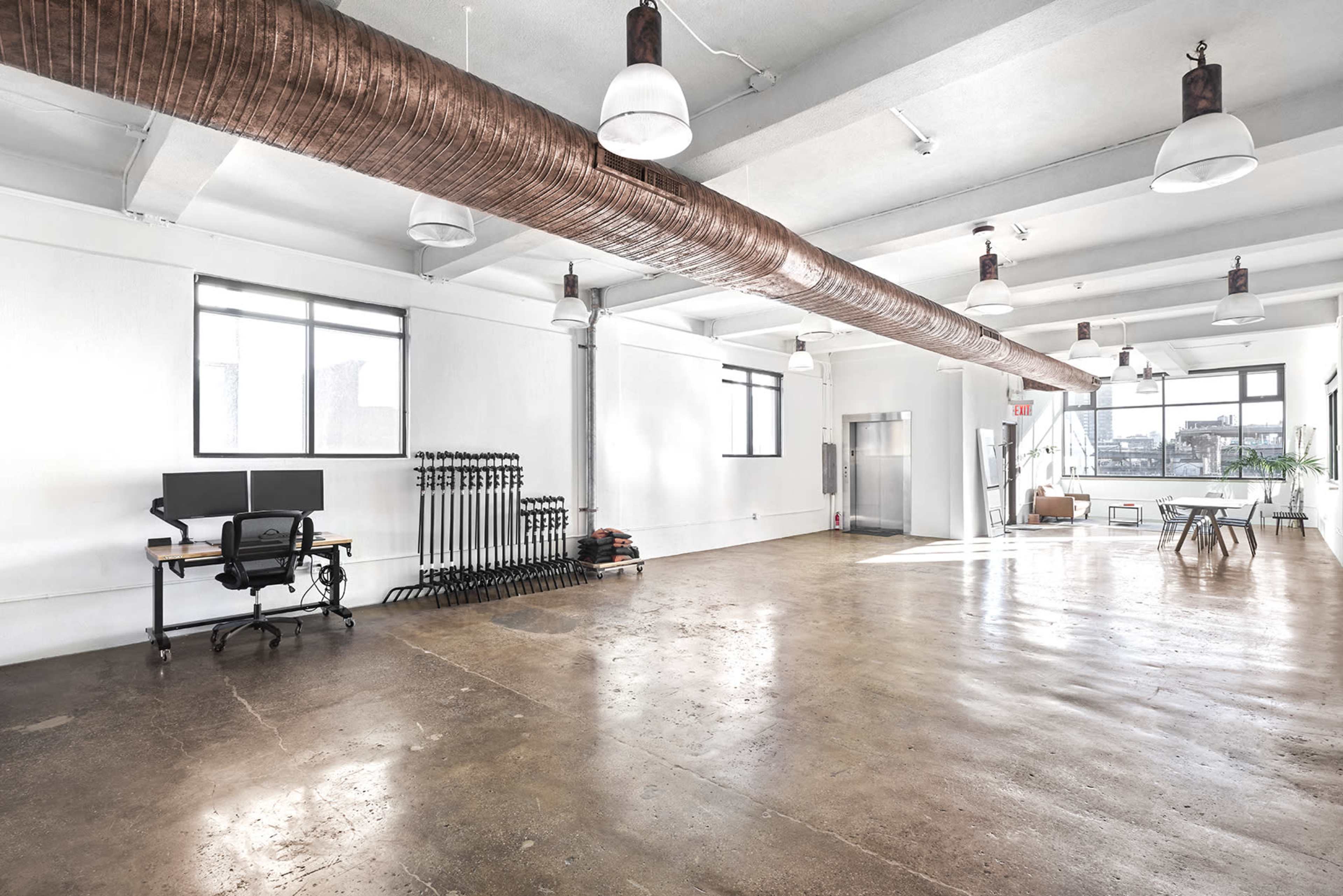 The image shows a spacious, empty room with concrete flooring, large windows, and a workstation featuring two computer monitors.