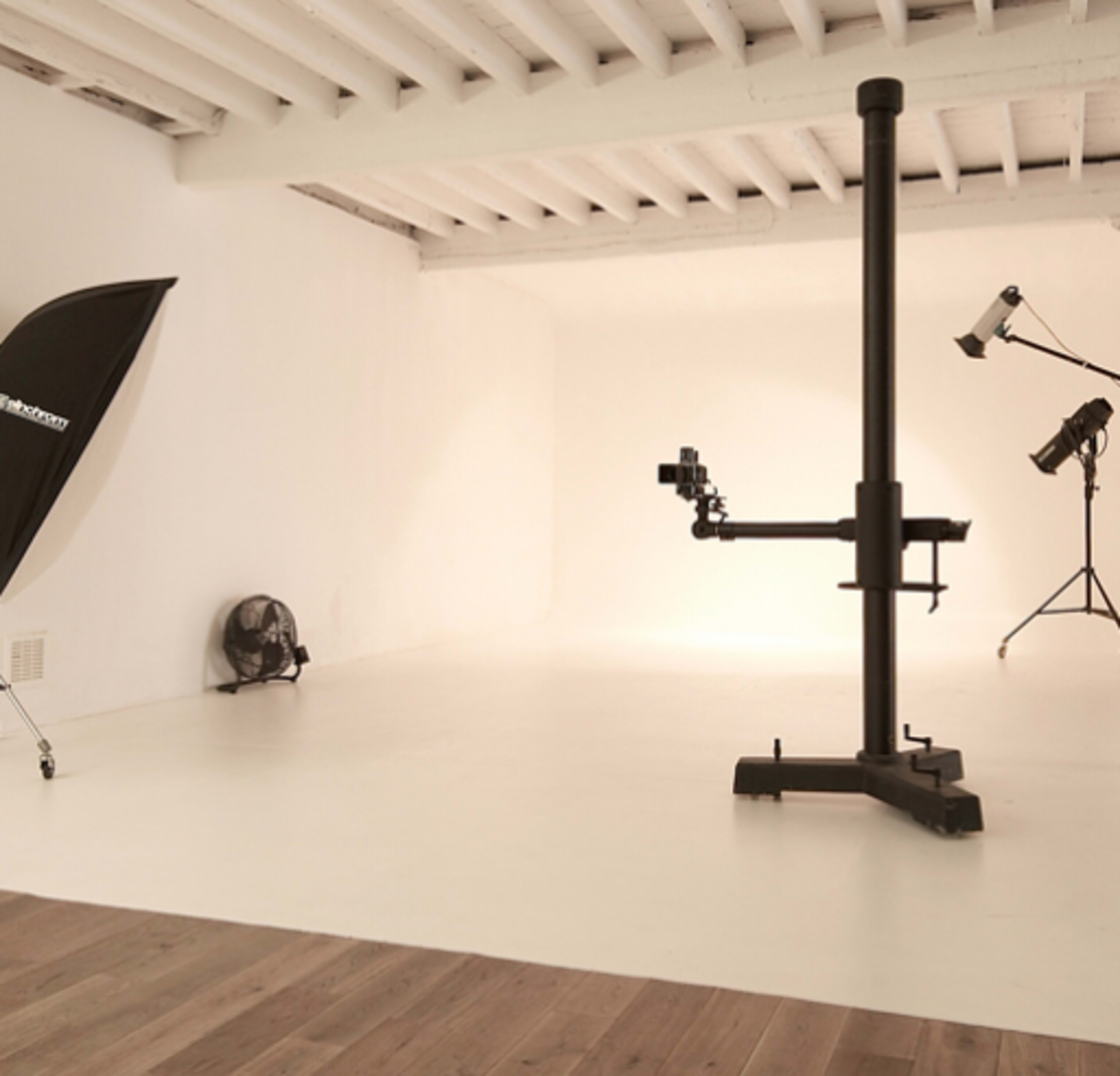 A minimalistic studio space with a white wall and floor, featuring a tripod with a light and a fan in the corner.