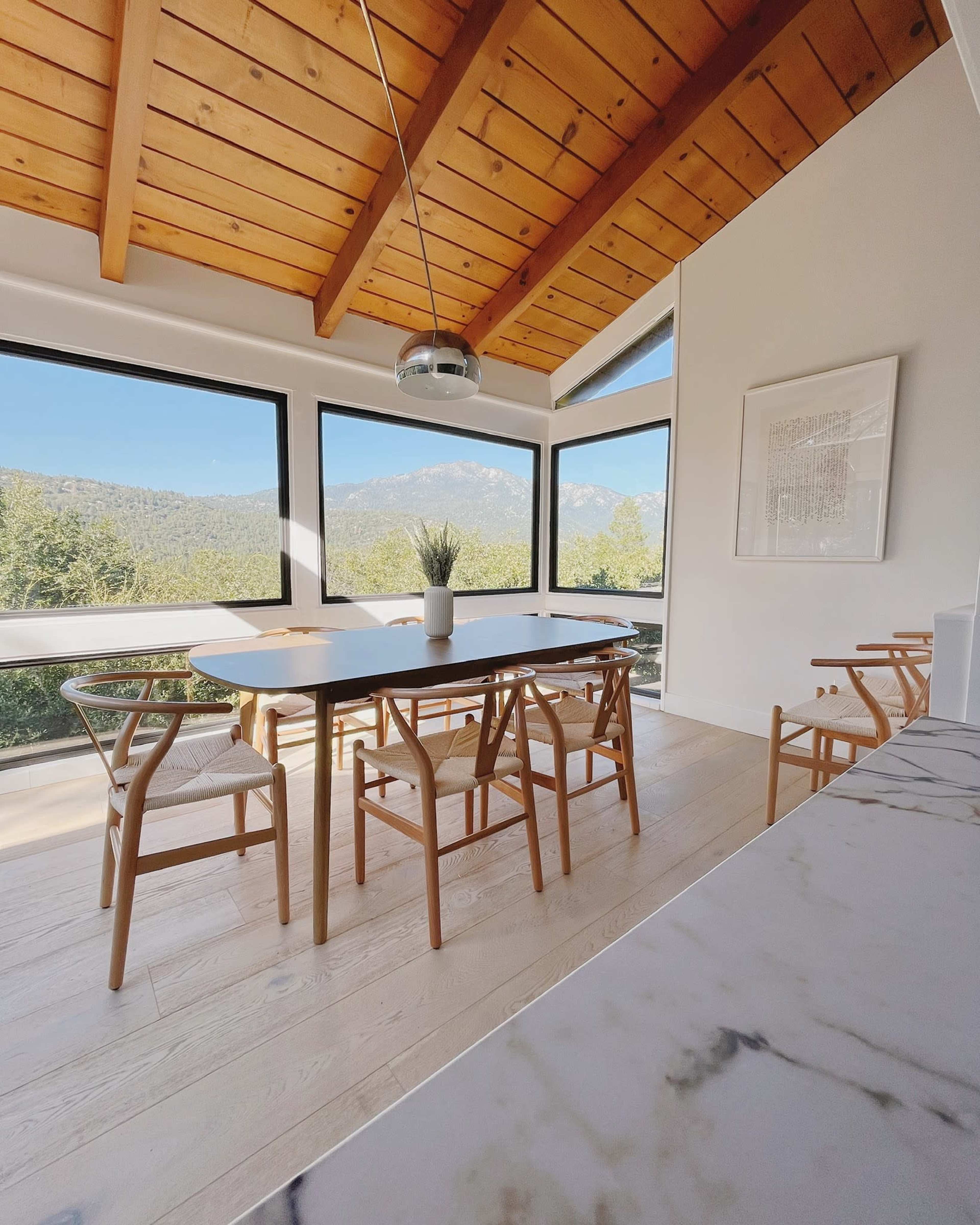 A dining area features a long white table surrounded by wooden chairs, with large windows showcasing a view of distant mountains.