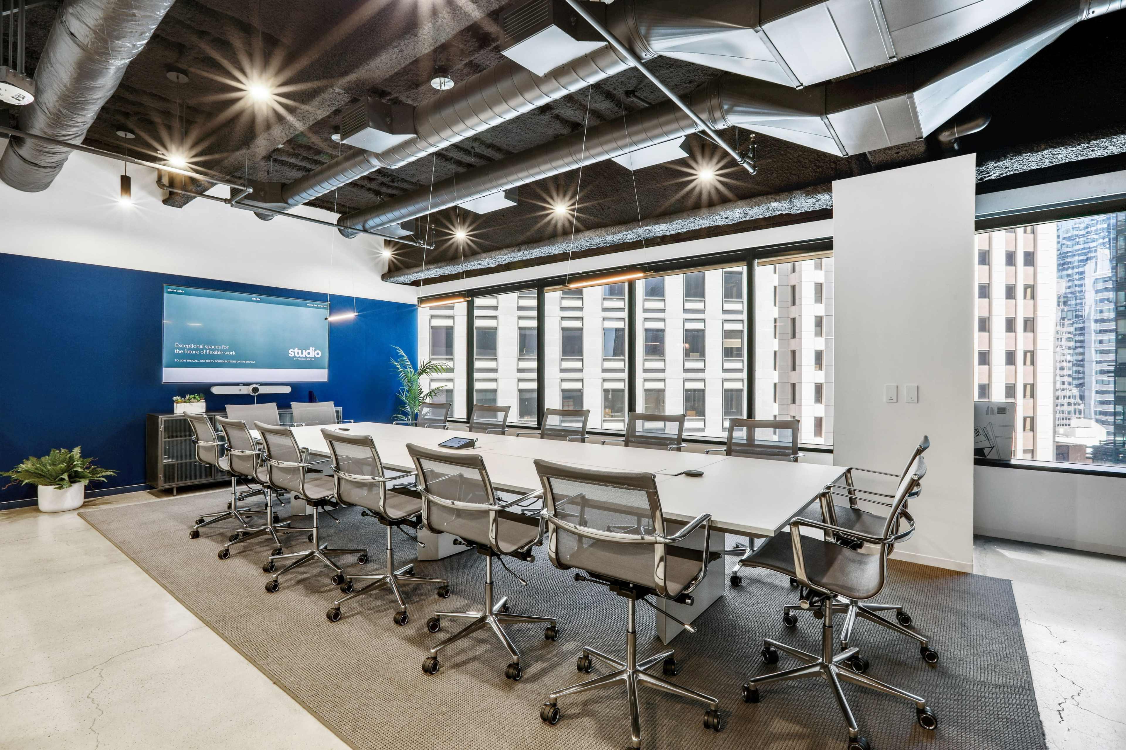A modern conference room features a long table surrounded by ergonomic chairs, with large windows providing views of a city skyline.