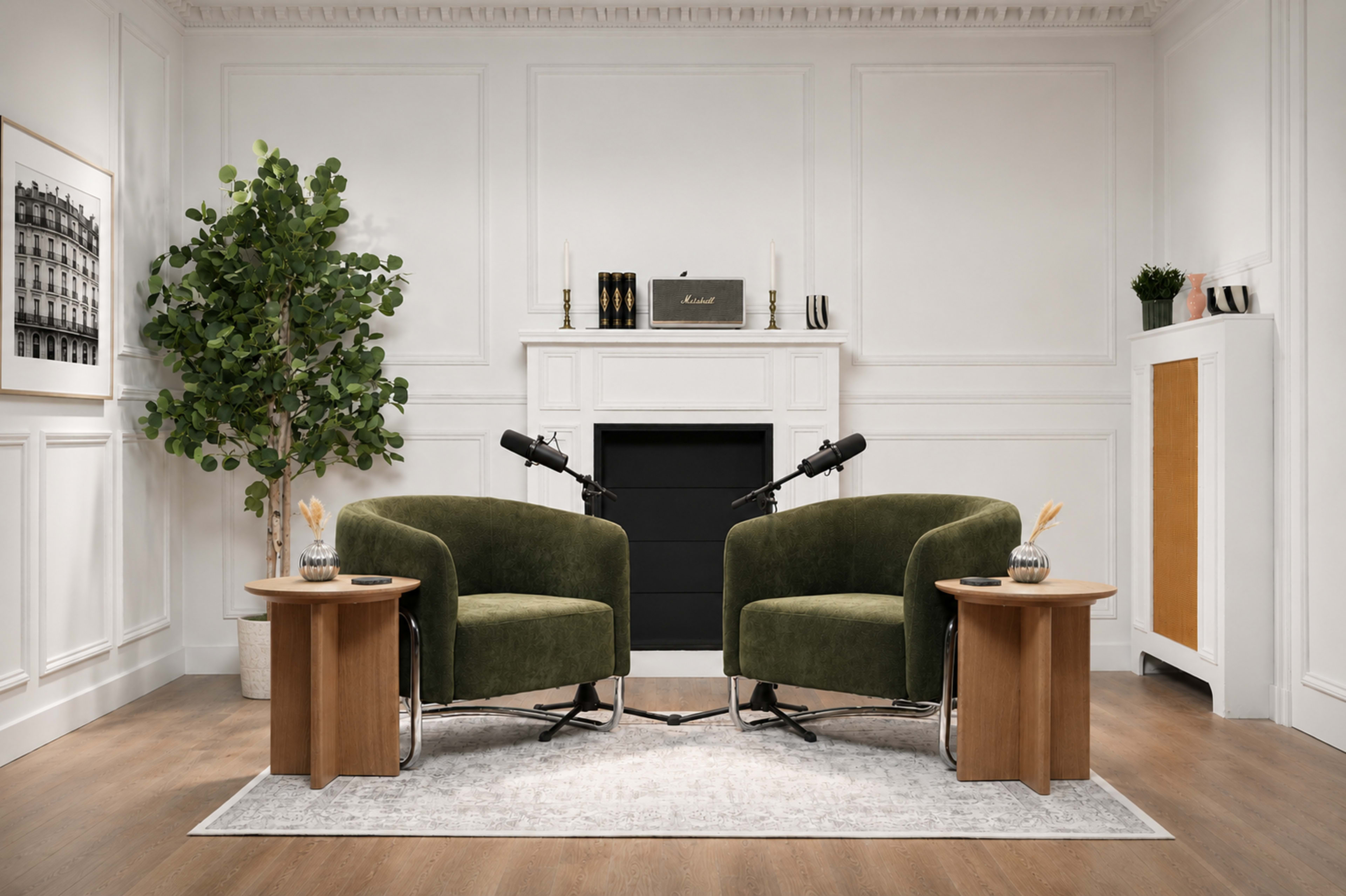 A minimalist interview setup features two green chairs facing each other with microphones on small wooden side tables, against a simple, elegant backdrop.