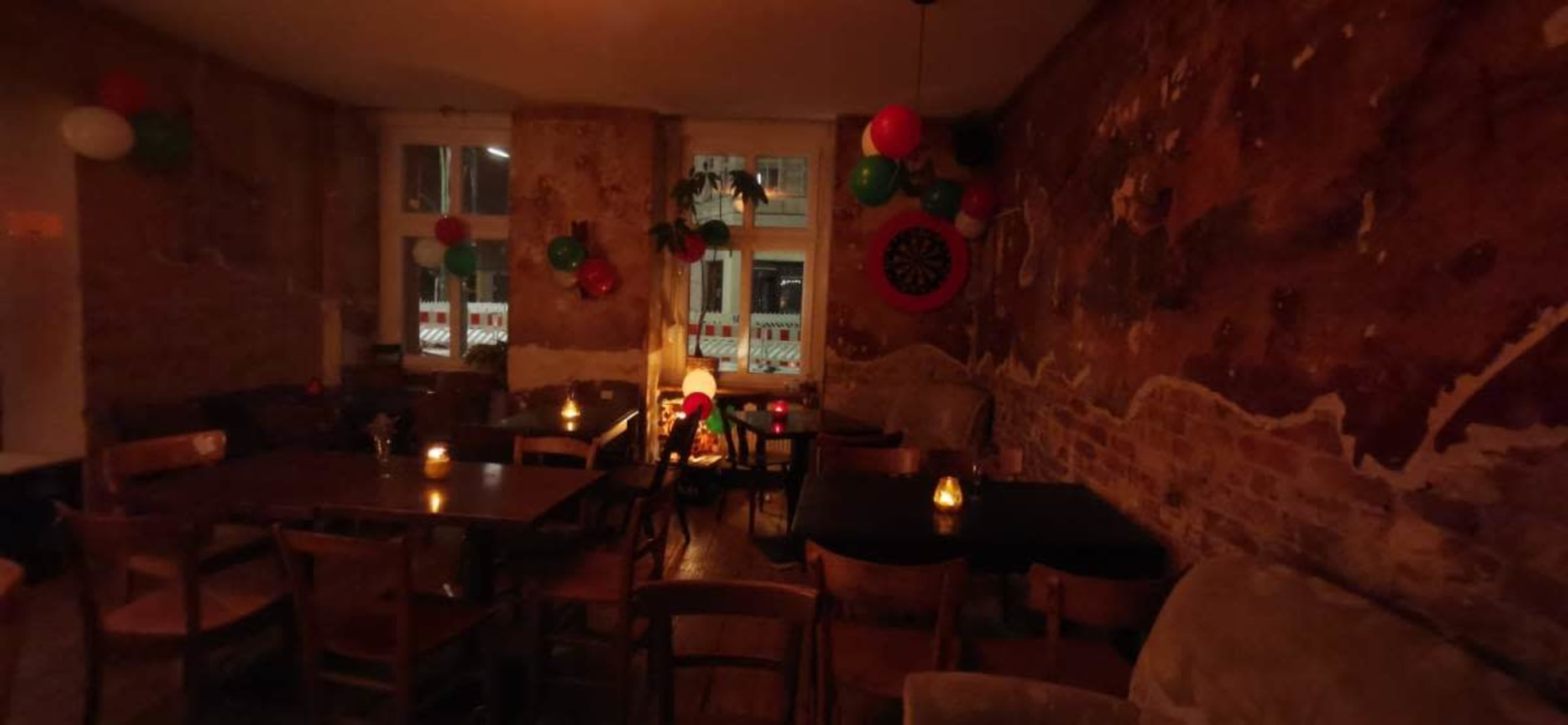 The image shows a dimly lit restaurant with exposed brick walls, wooden tables, and colorful decorations, including balloons and a dartboard.