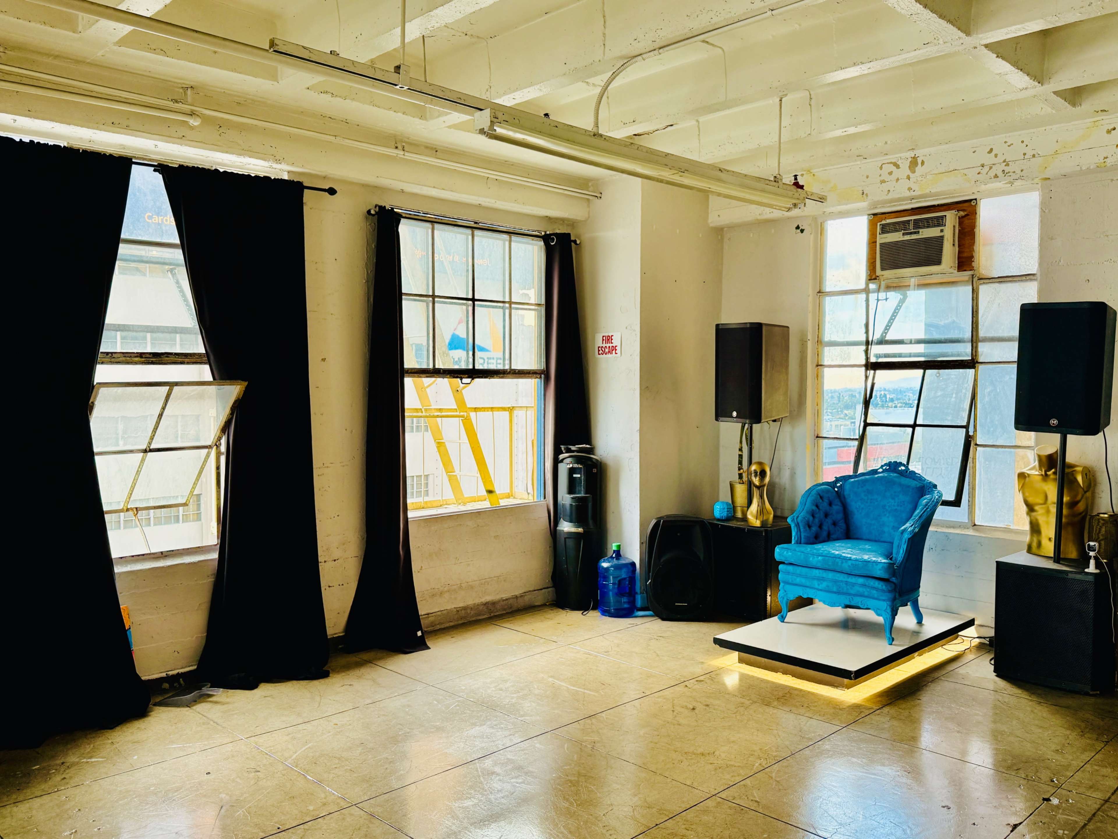 The image shows a brightly lit room with two large windows, black curtains, a blue chair on a platform, and two speakers positioned on either side.
