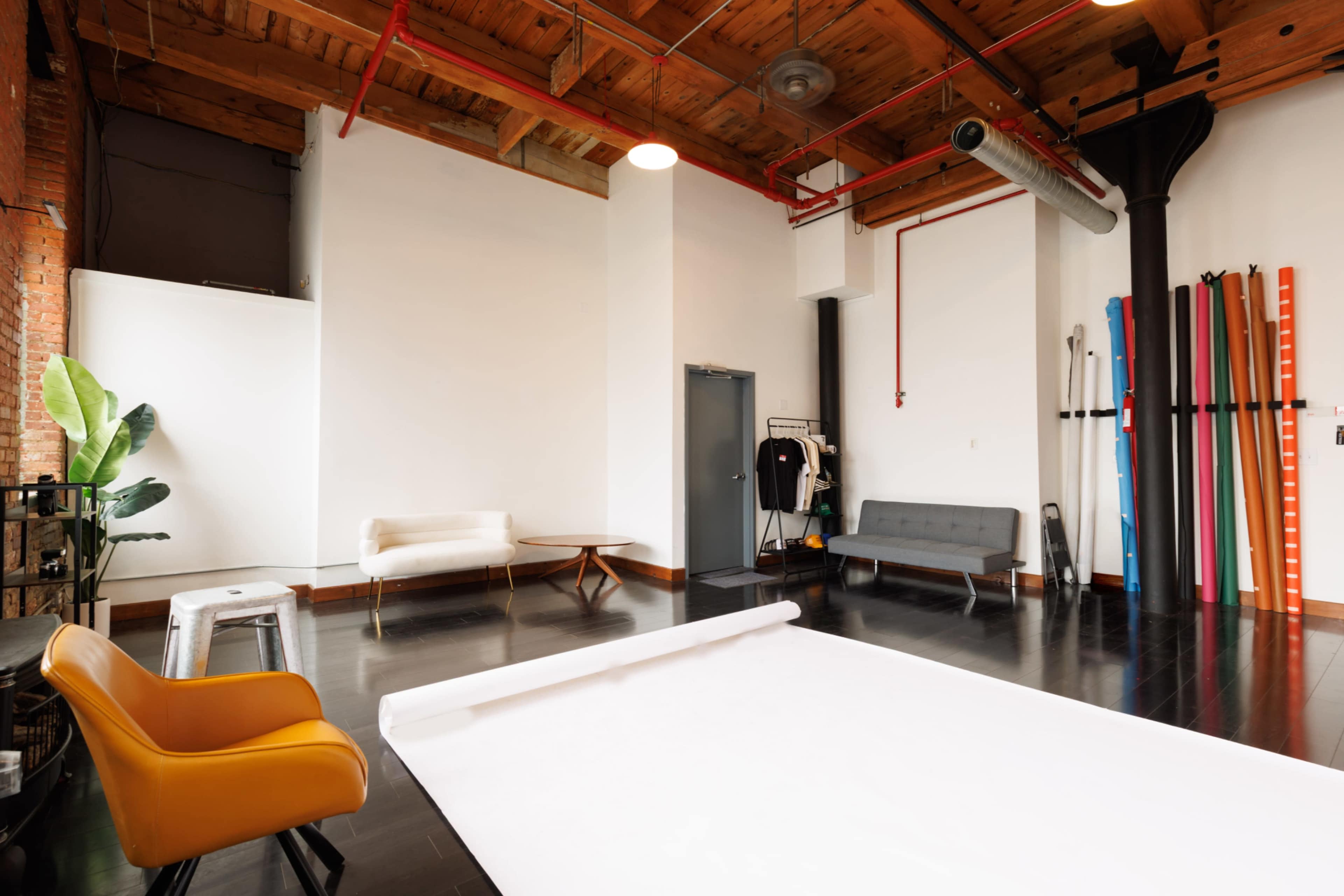 The image shows a spacious, well-lit studio with wooden ceilings, hardwood floors, and minimalist furniture, including a sofa, a table, and a chair.