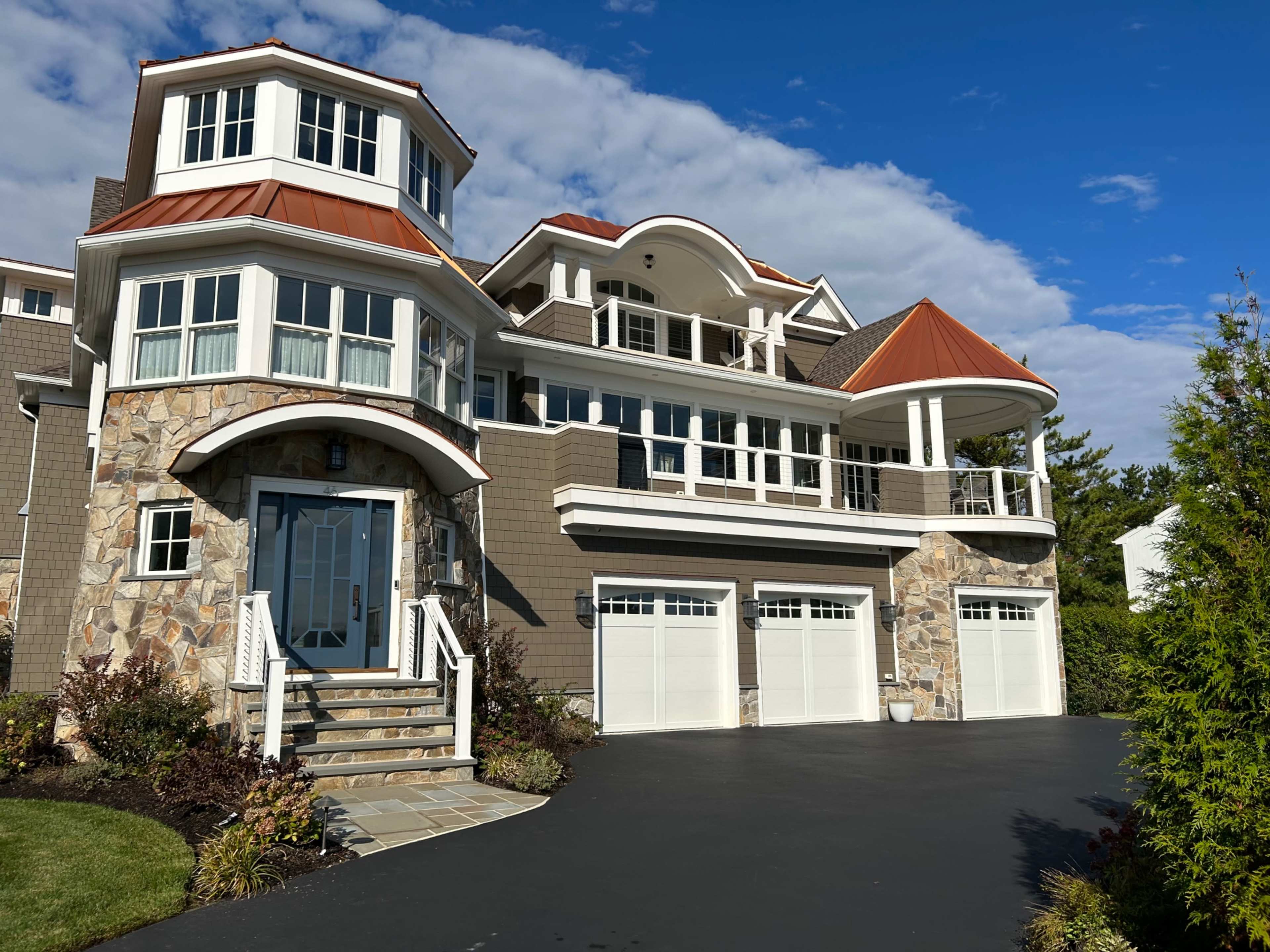The image showcases a large two-story house with a stone exterior, multiple gabled roofs, and a landscaped driveway leading to three garage doors.