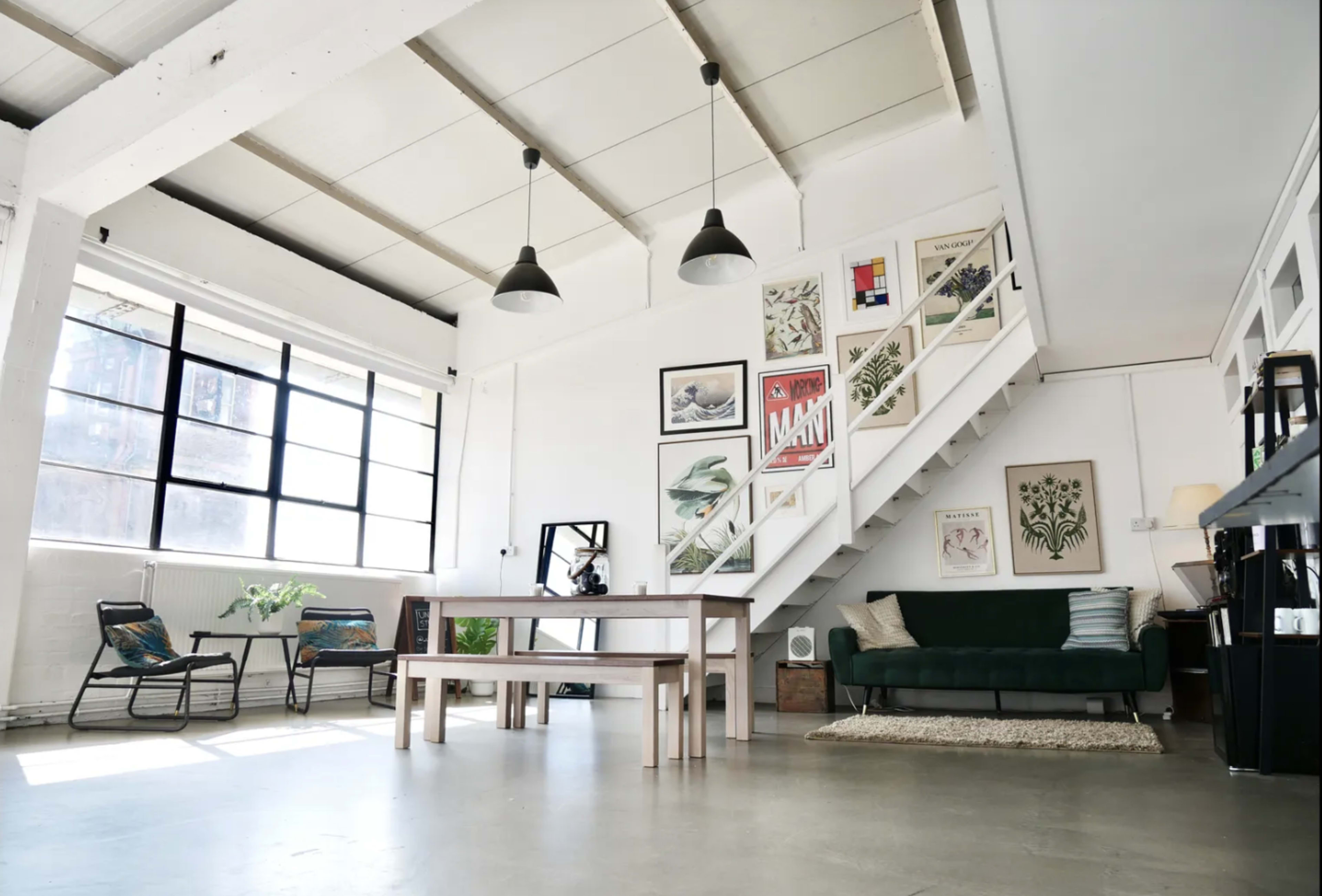The image shows a spacious modern interior with a staircase, a dining table, seating area, and large windows allowing natural light to enter.