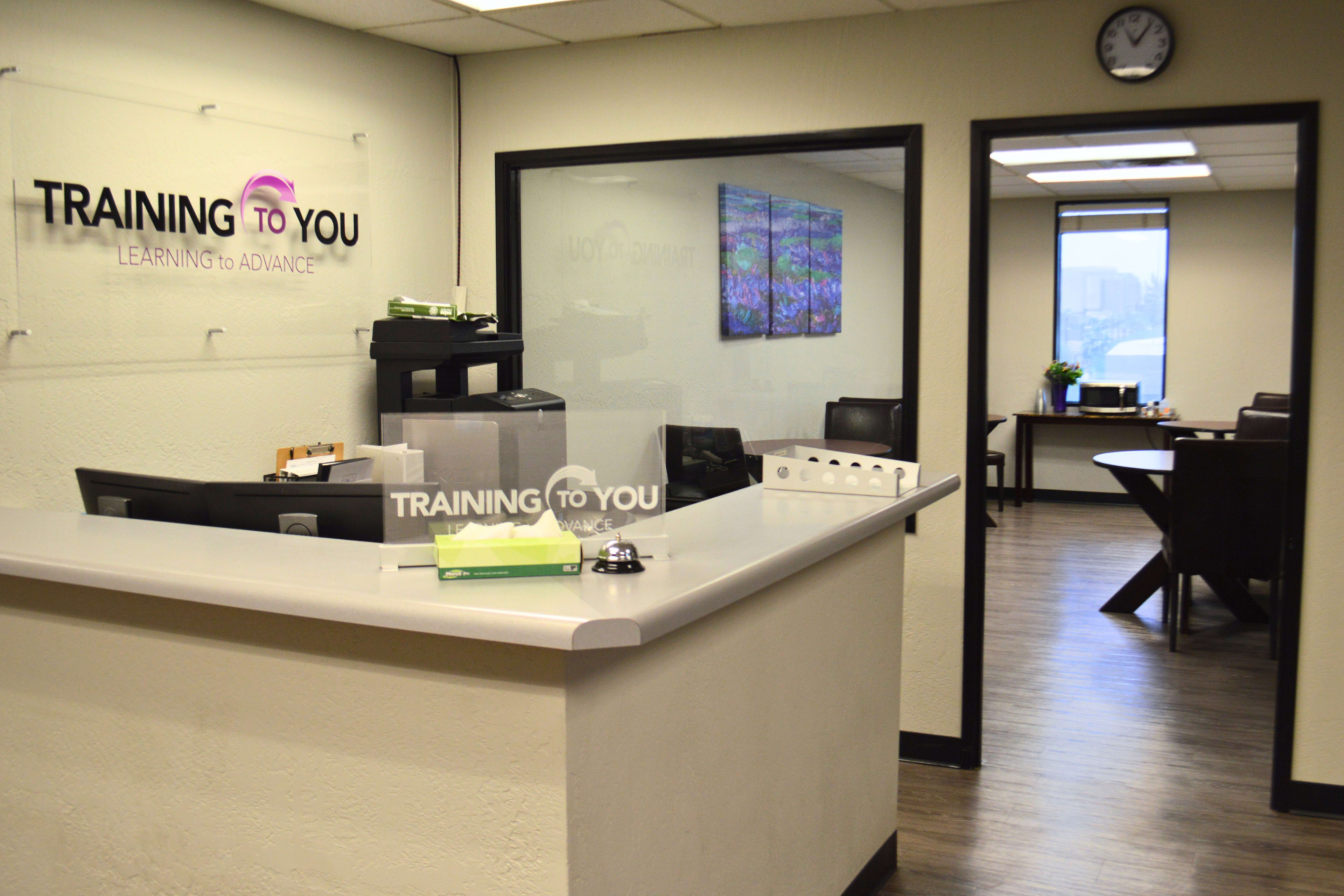 The image shows a reception area with a gray counter and a glass wall leading to a conference room, featuring a logo that reads "TRAINING TO YOU."