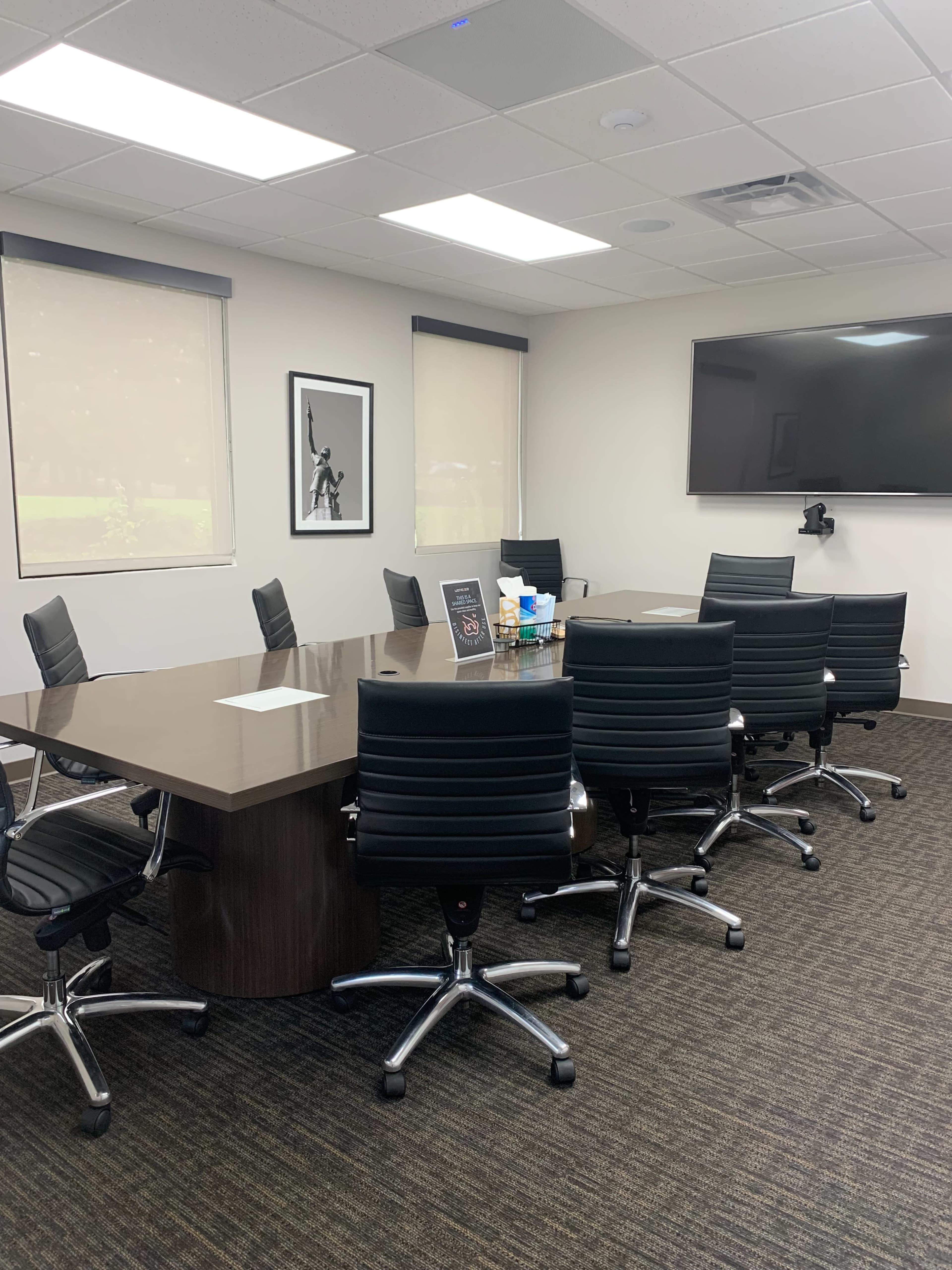 A modern conference room features a long wooden table surrounded by black office chairs, with a large screen on one wall and windows covered by blinds.