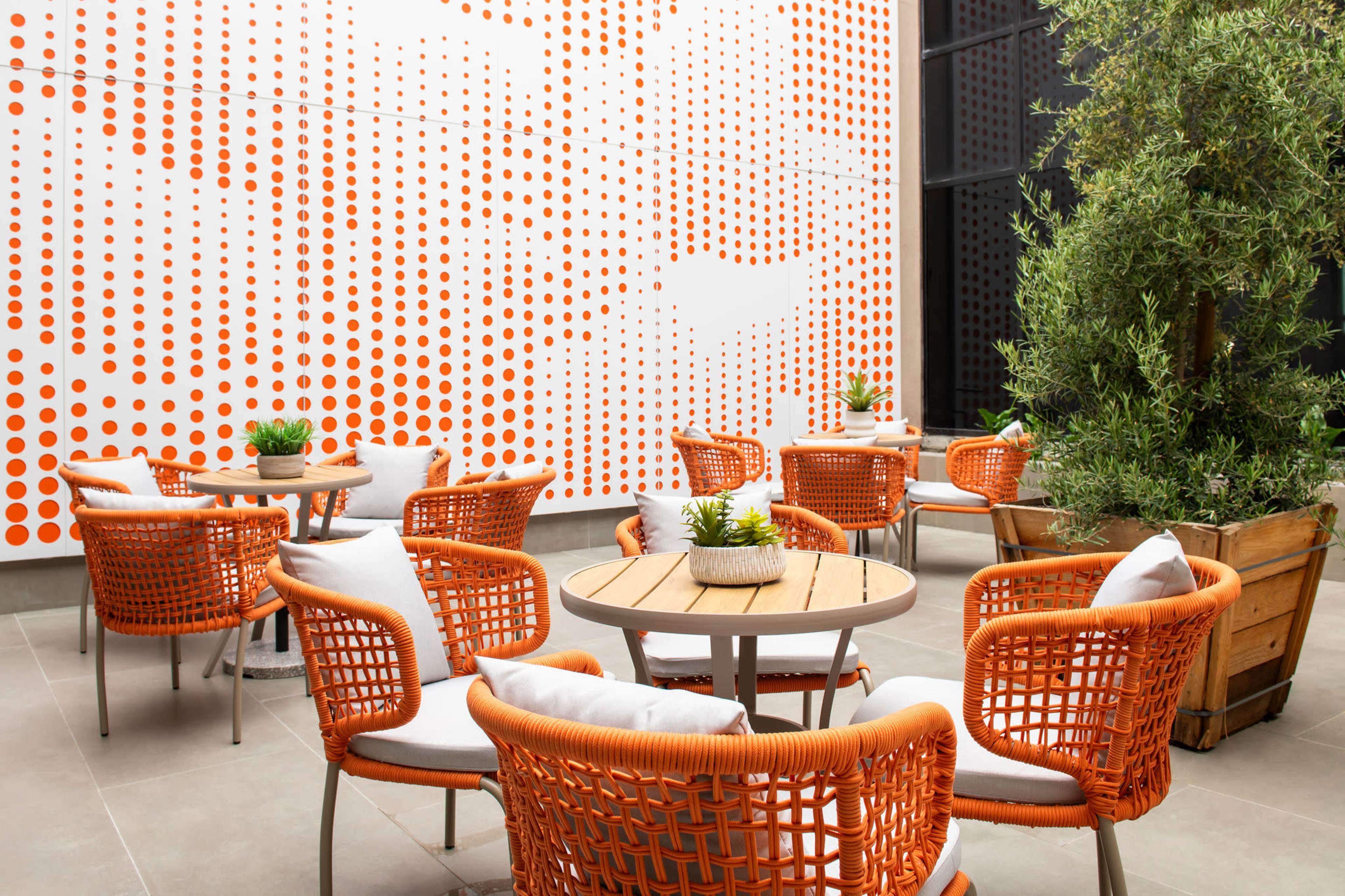 A patio features several round tables surrounded by orange woven chairs, with a backdrop of a dotted orange wall and a potted plant.