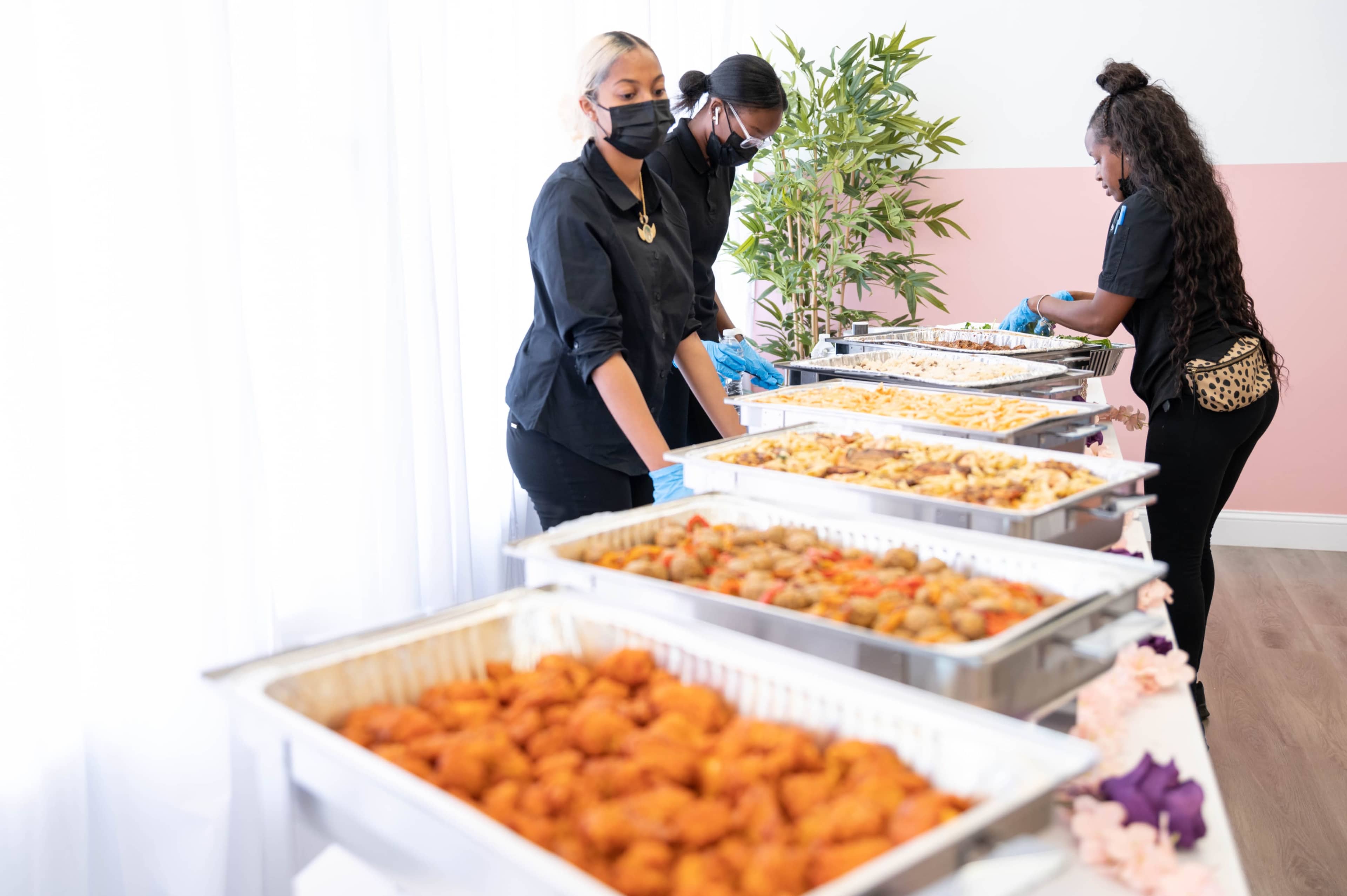 Caterers serve a variety of food from metal trays at a buffet-style setup in a decorated room.