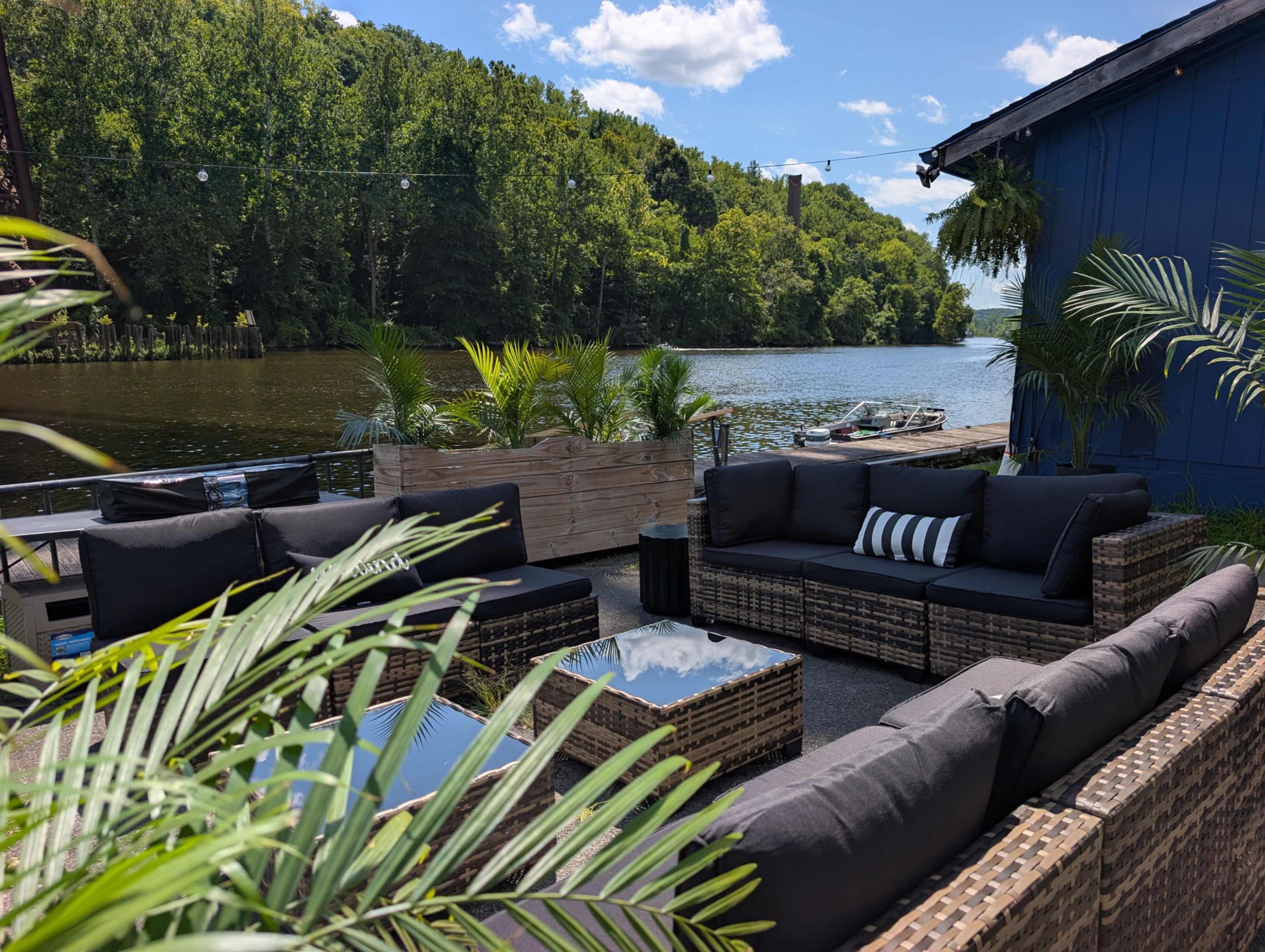 The image shows a riverside outdoor seating area with wicker furniture, potted plants, and a view of trees and water in the background.