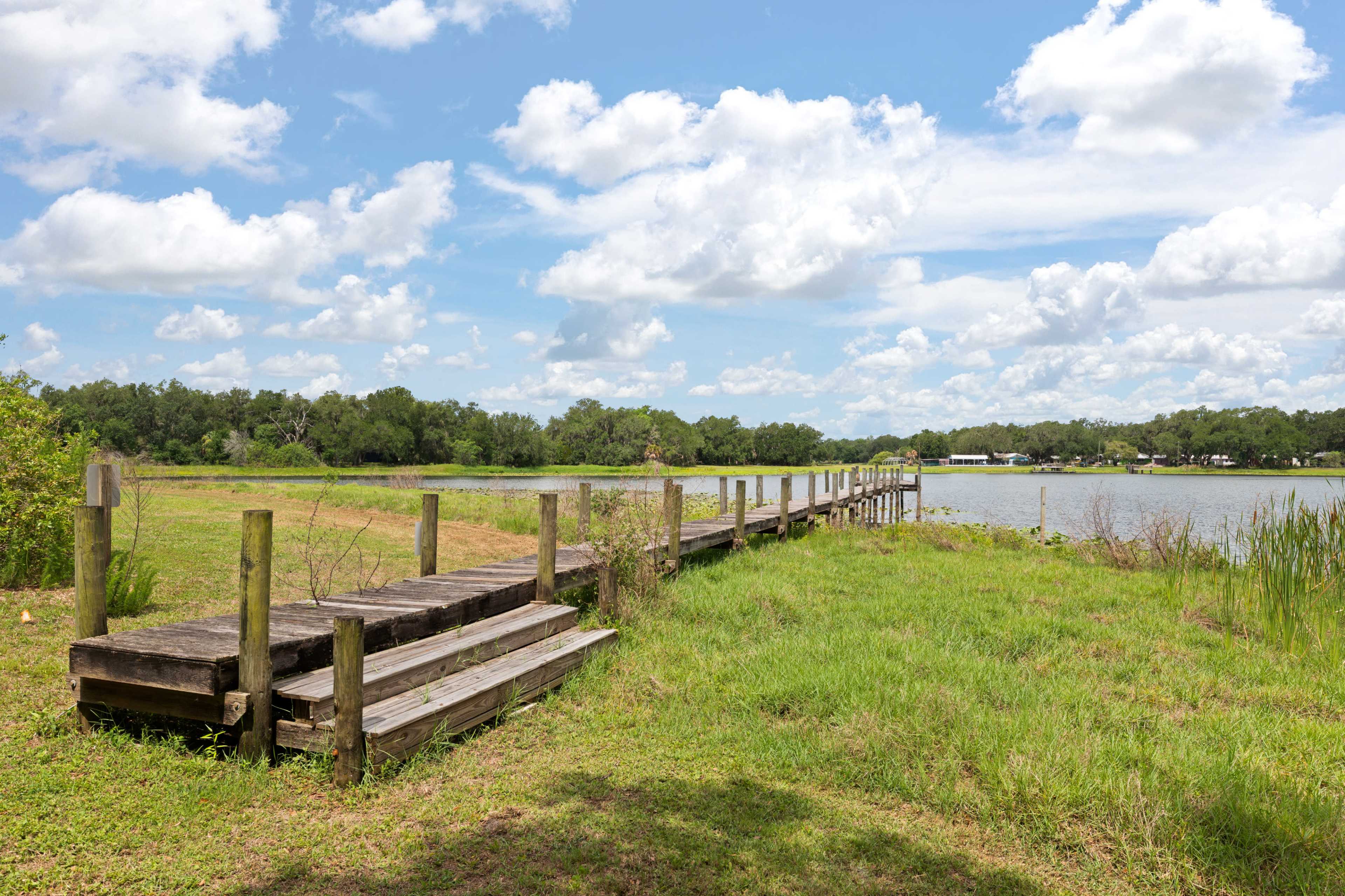 A wooden dock extends into a calm body of water, surrounded by lush greenery and blue skies with scattered clouds.