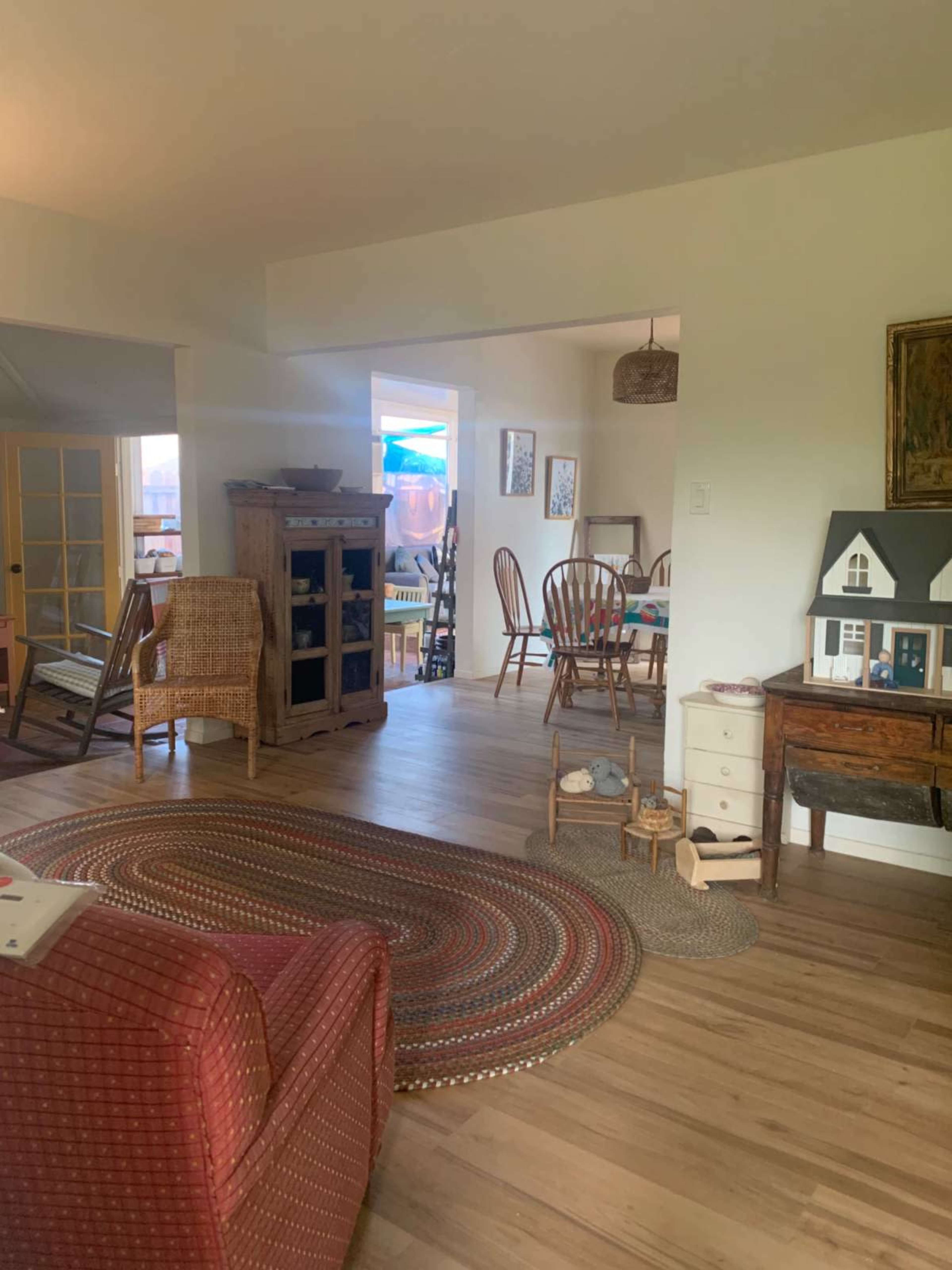 The image shows a cozy interior space with a living area featuring a red chair, a wooden cabinet, and a rug, leading into a dining area with a wooden table and chairs.