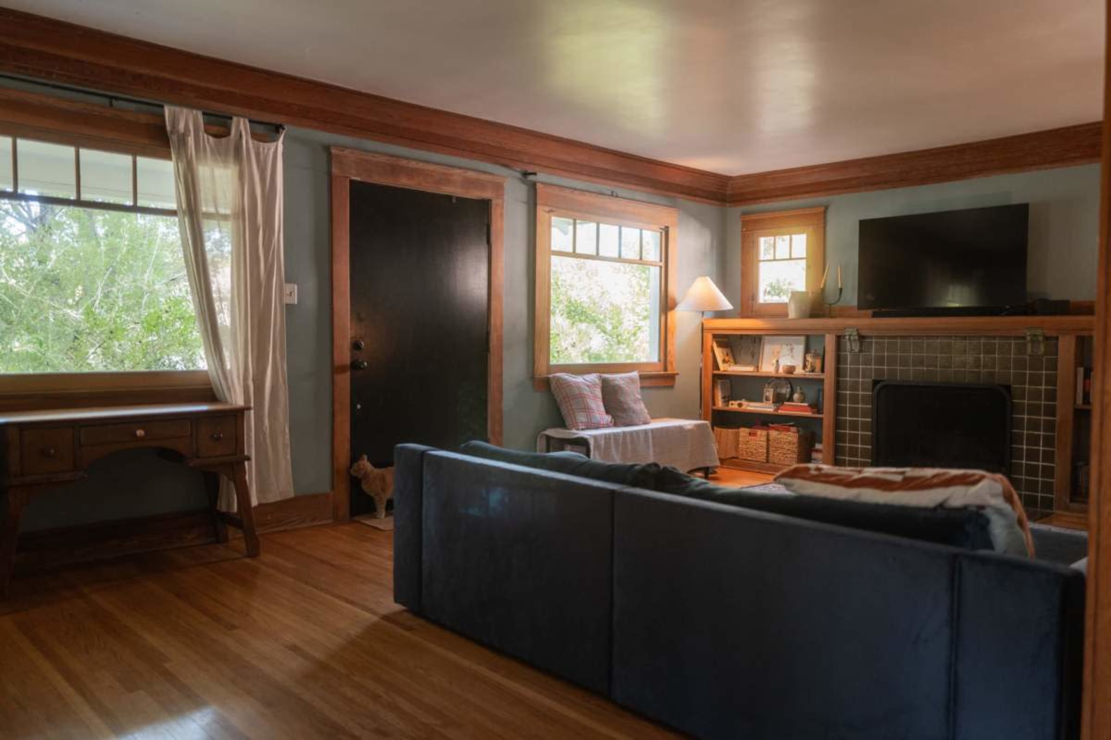 The photo shows a living room with a blue couch, a wooden desk, a flat-screen TV above a fireplace, and windows that let in natural light.
