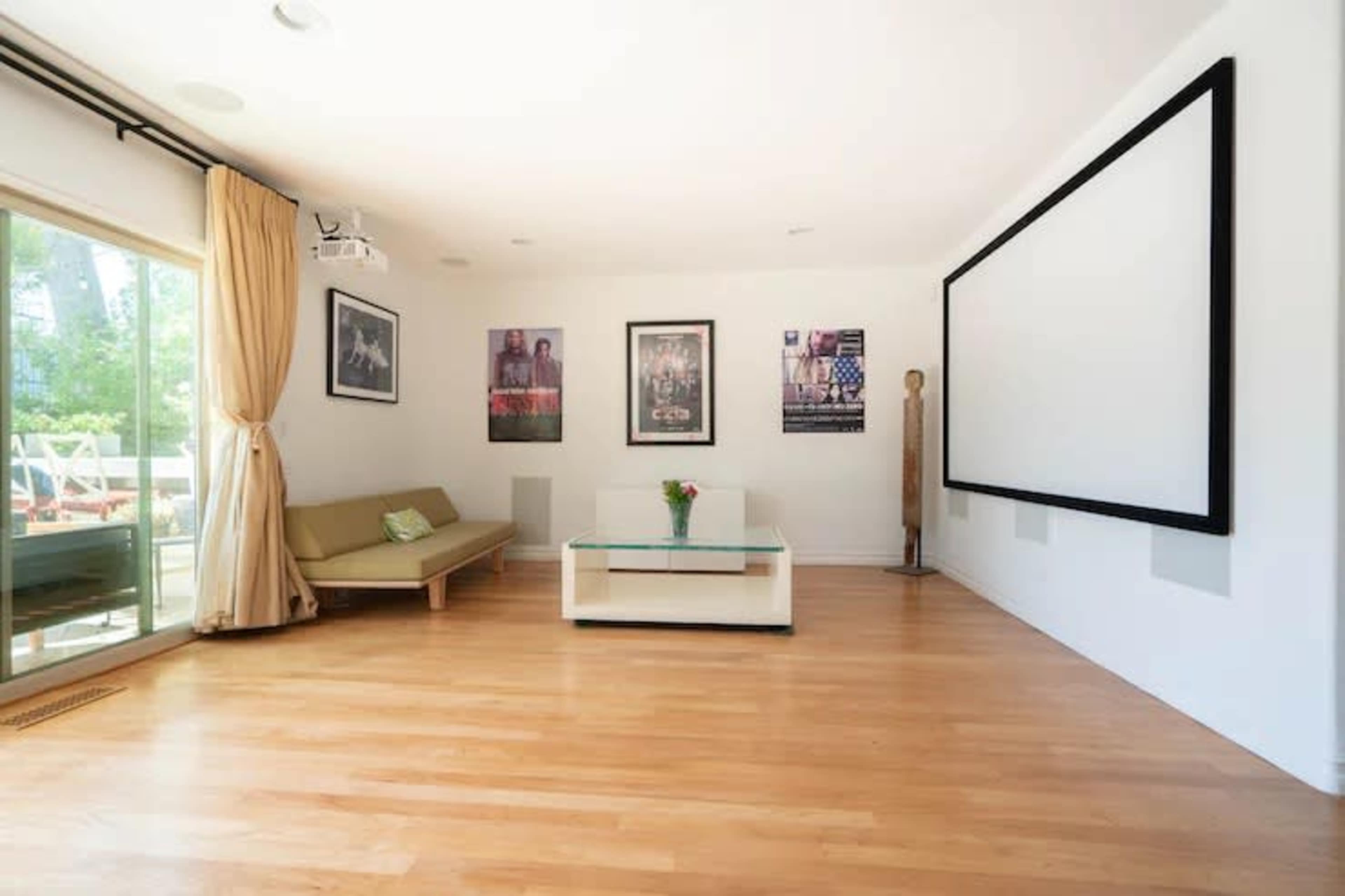 A bright room features a large projector screen, a coffee table with a vase of flowers, and posters on the walls.