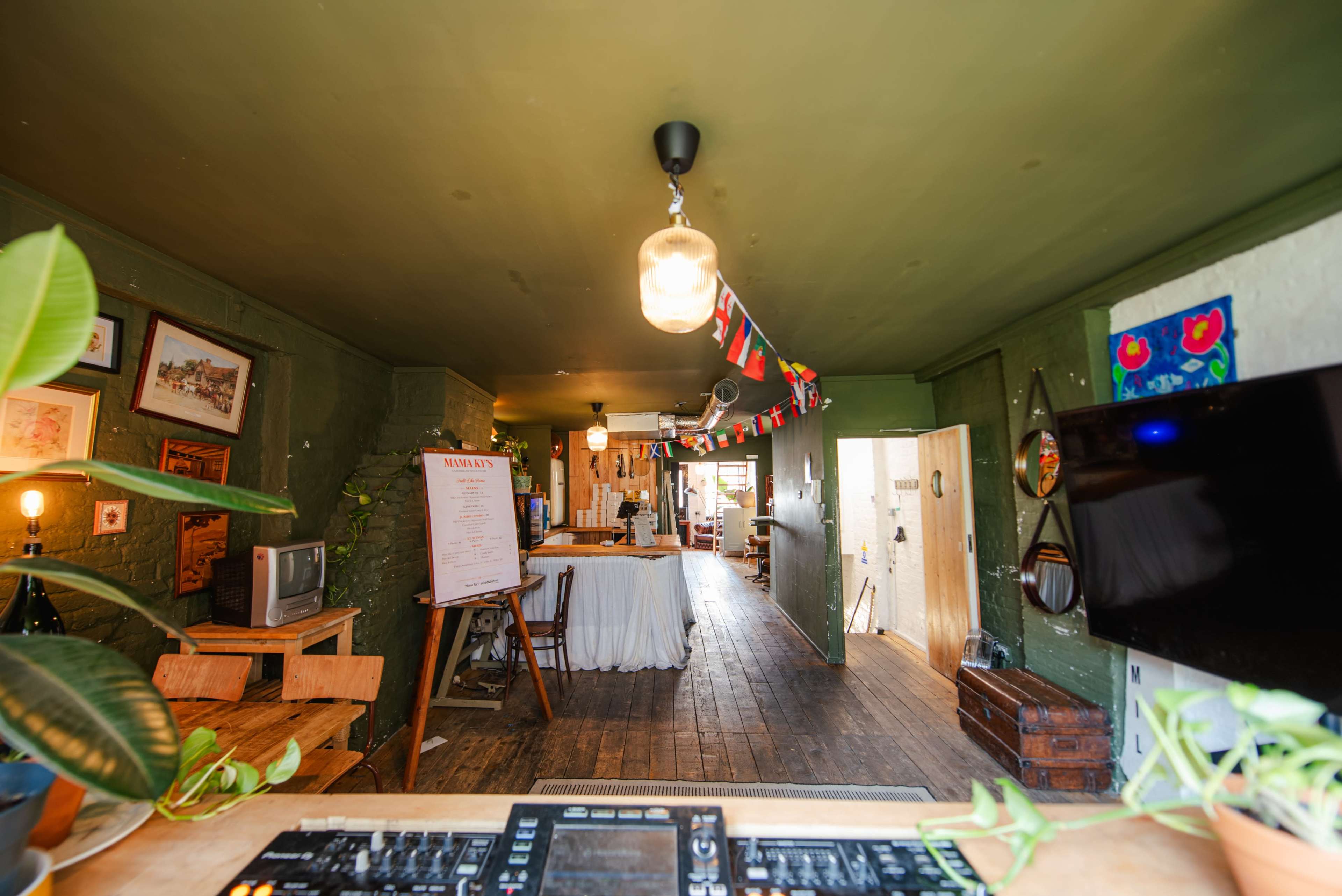 The image shows the interior of a cozy café or restaurant featuring wooden flooring, decorative plants, and an inviting layout with tables and colorful bunting.