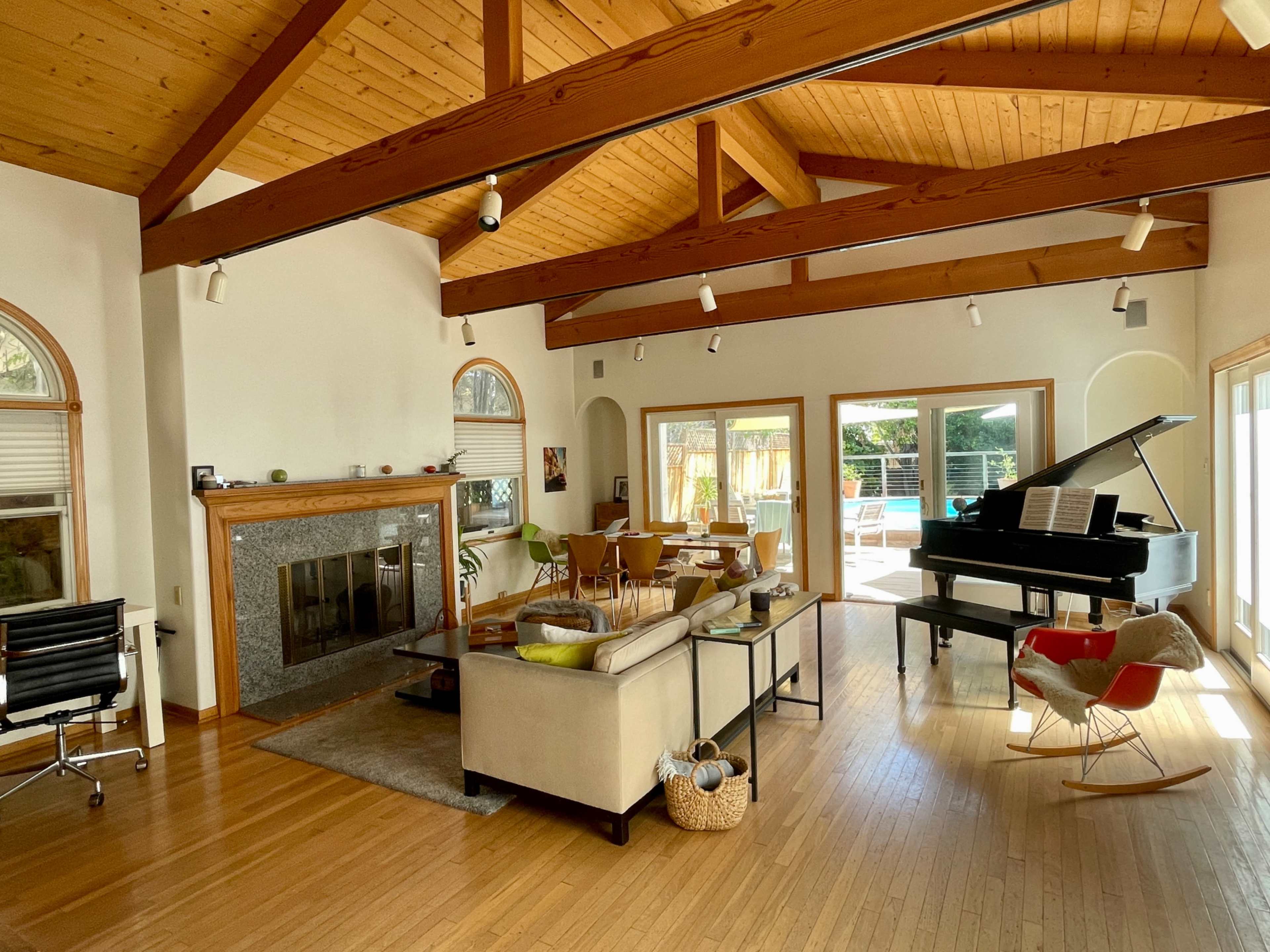 The image shows a spacious living room with a grand piano, a fireplace, large windows, and direct access to an outdoor pool area.