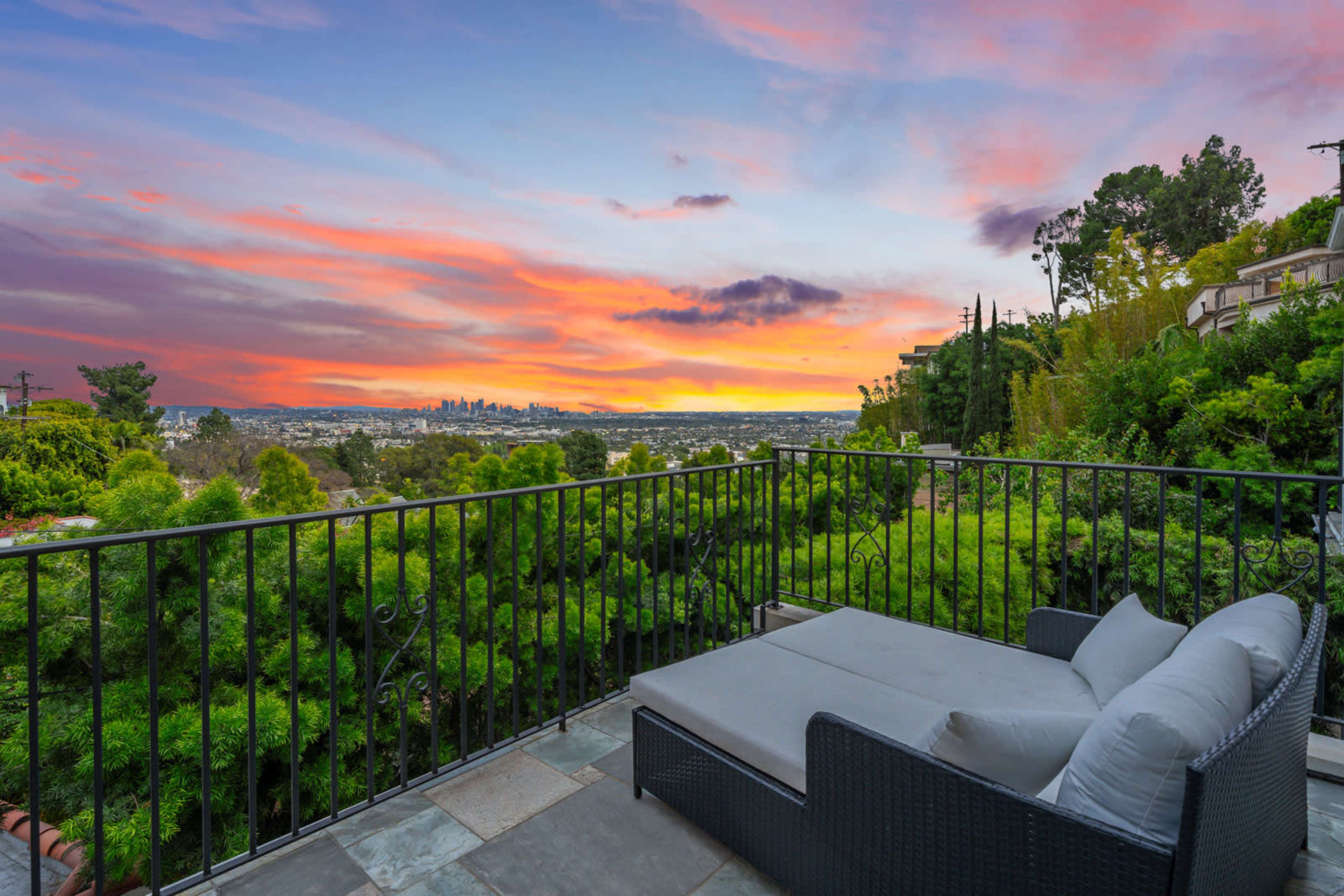 A balcony overlooking a city skyline at sunset, framed by lush greenery.