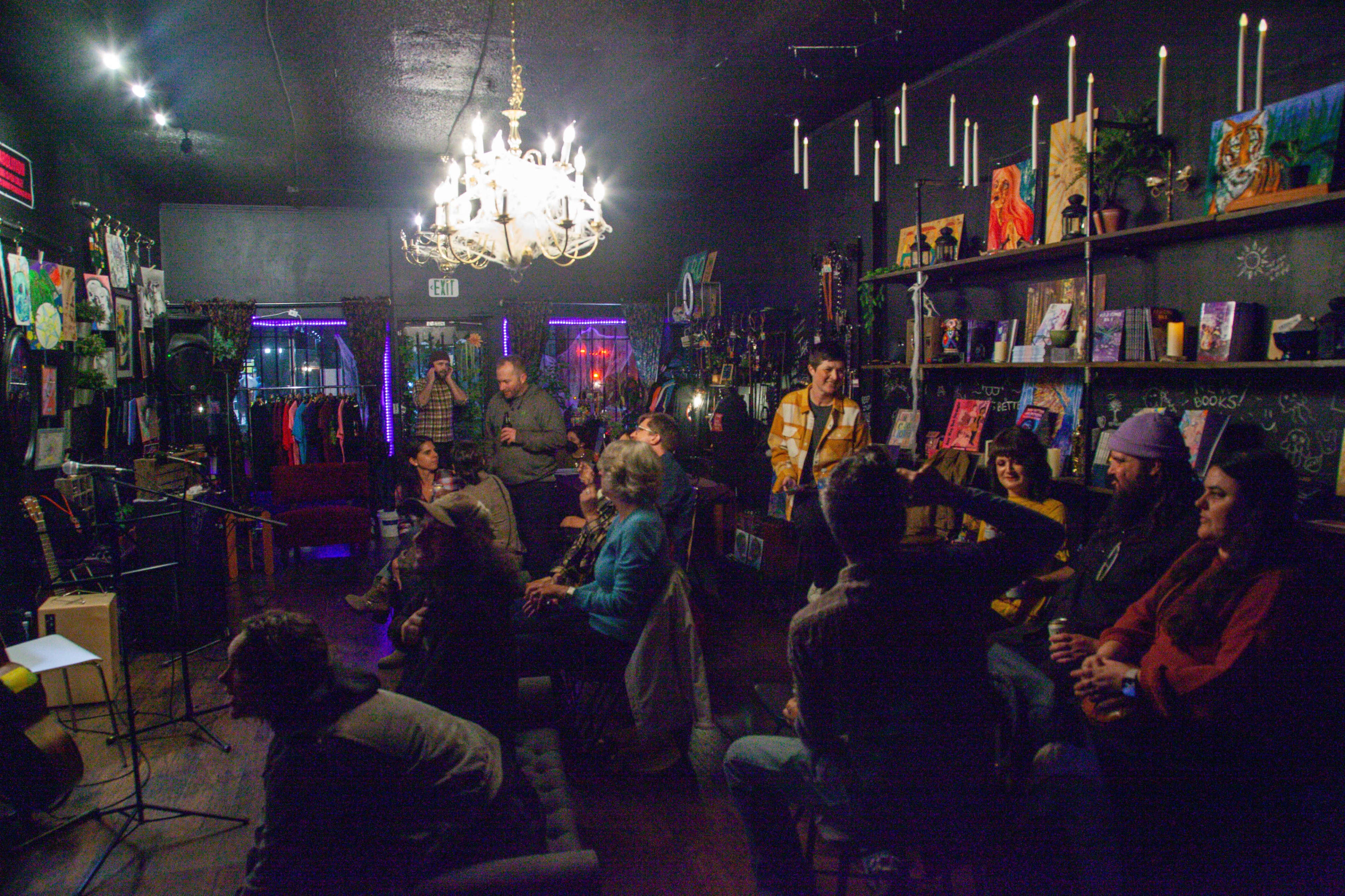 A group of people sits in a cozy room filled with artwork and a chandelier, engaged in a performance or gathering.