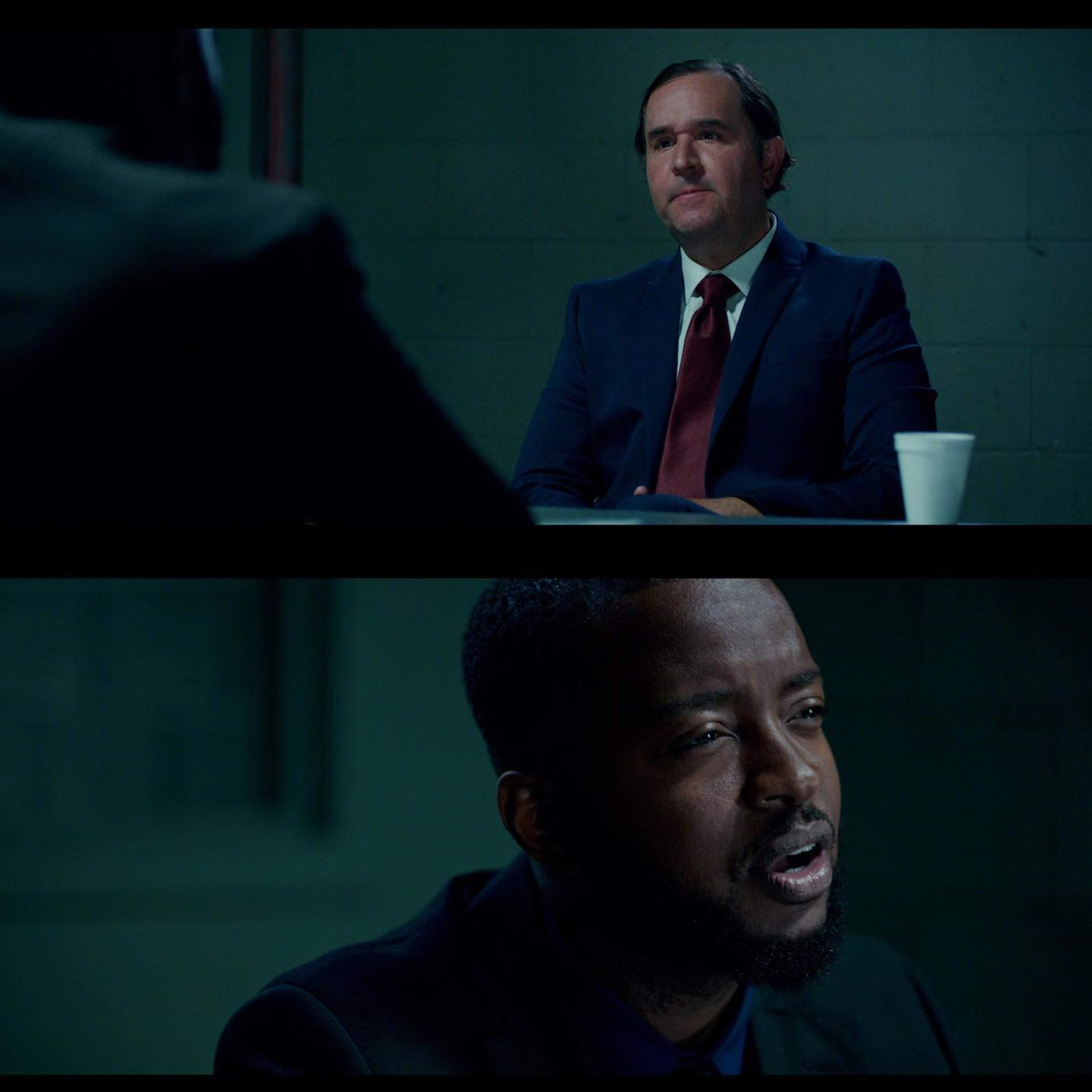 A man in a suit sits at a table across from another man, both engaged in a serious conversation in a dimly lit room.