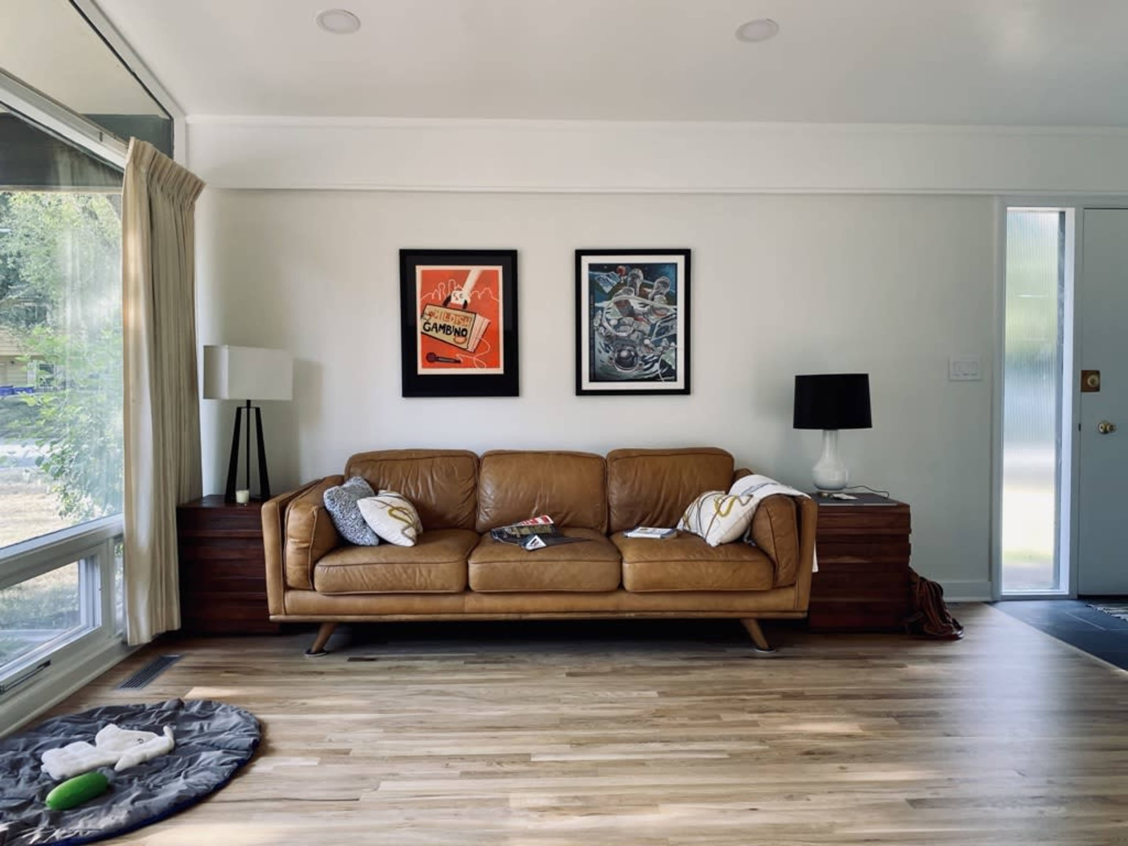 The image shows a living room featuring a brown leather sofa, two framed artworks on the wall, a side table with a lamp, and a wooden cabinet against the wall.