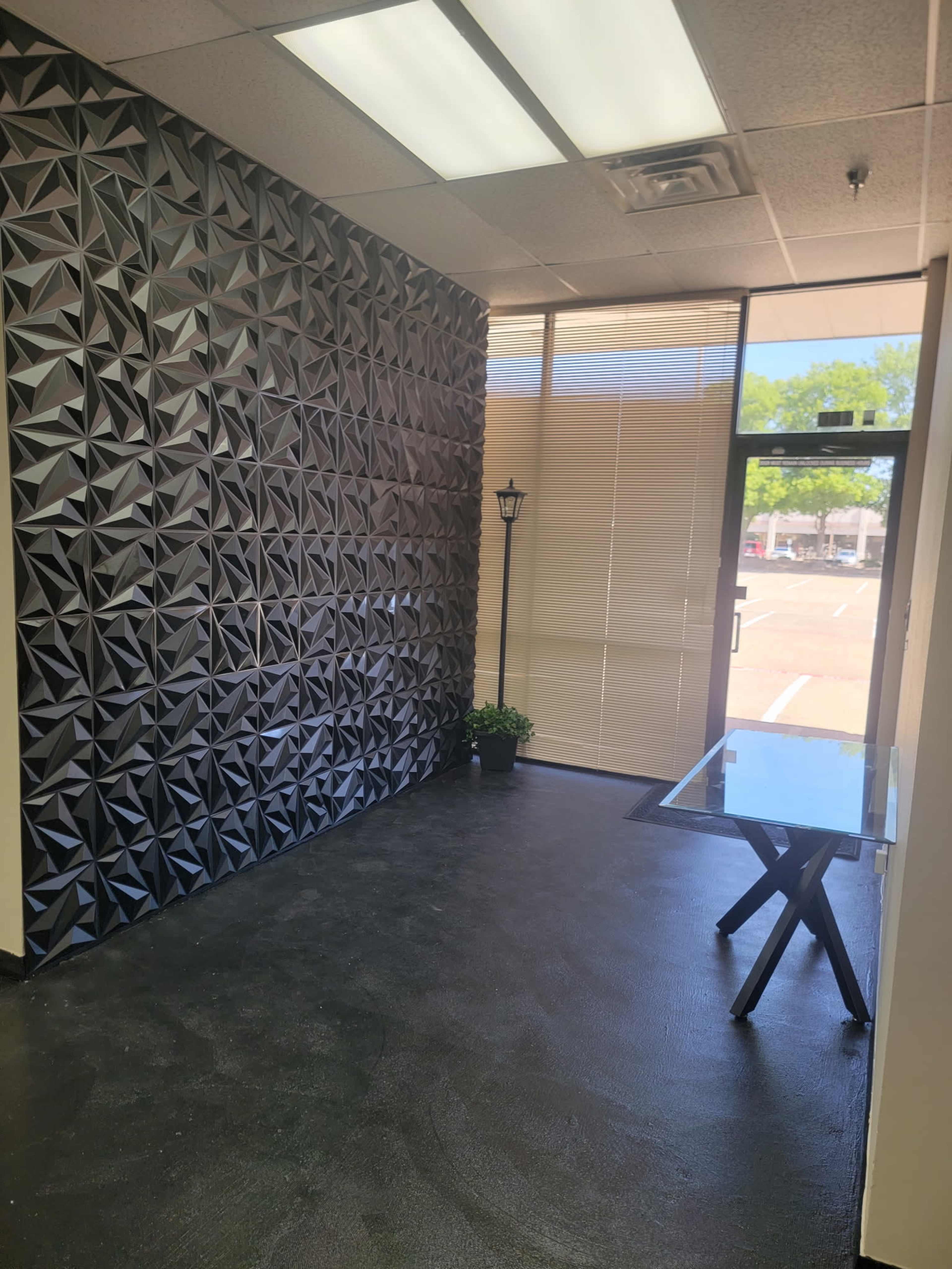 The image shows a small reception area featuring a textured black wall, a glass table, and a doorway with blinds leading outside.