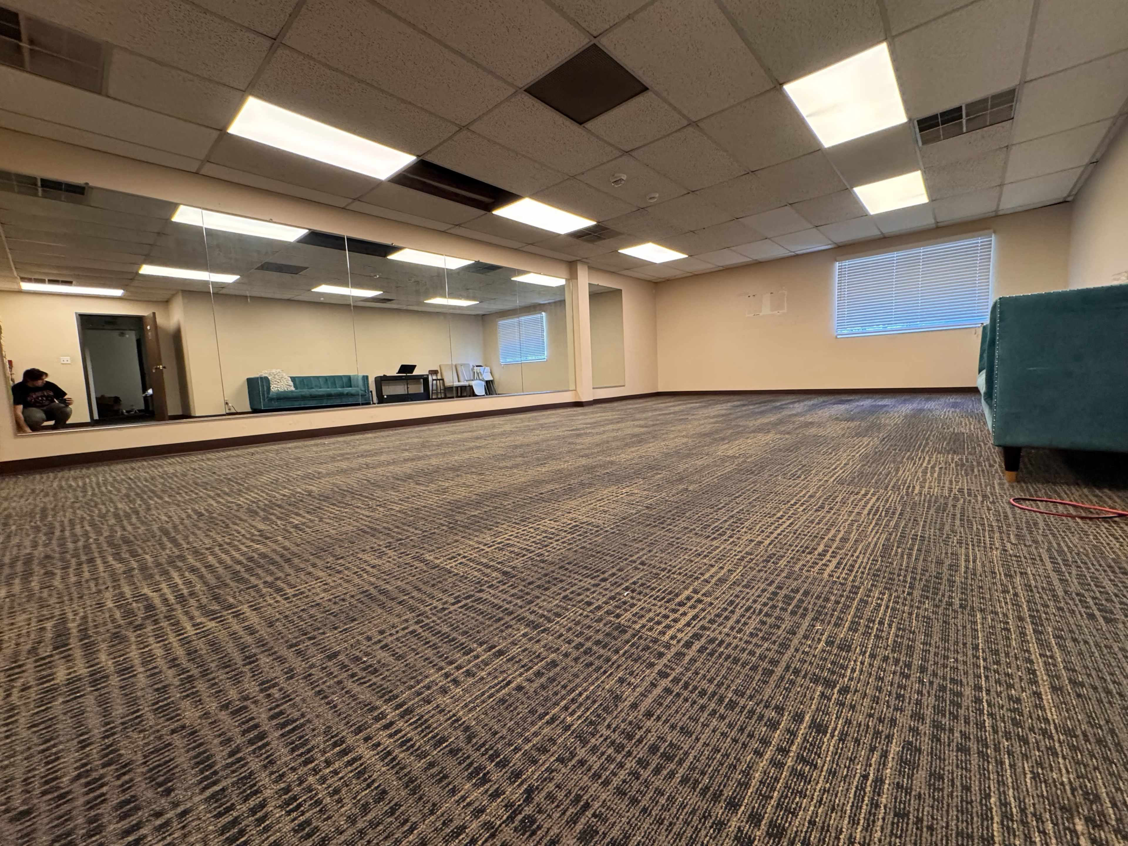 The image shows a spacious, empty room with carpeted flooring, a large mirror on one wall, and a few pieces of furniture in the corner.