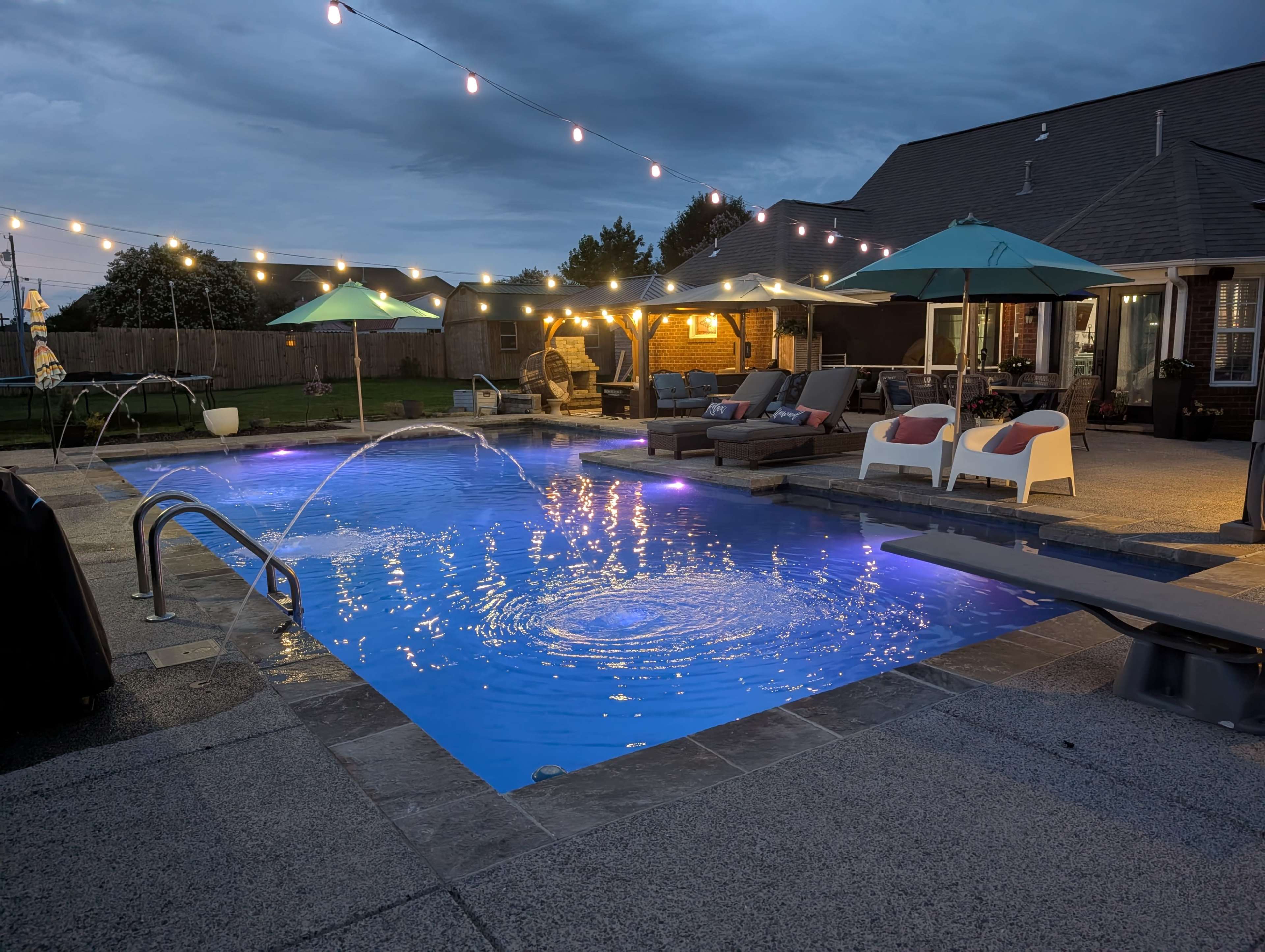 The image shows a well-lit backyard pool area with lounge furniture, water features, and umbrellas under a twilight sky.
