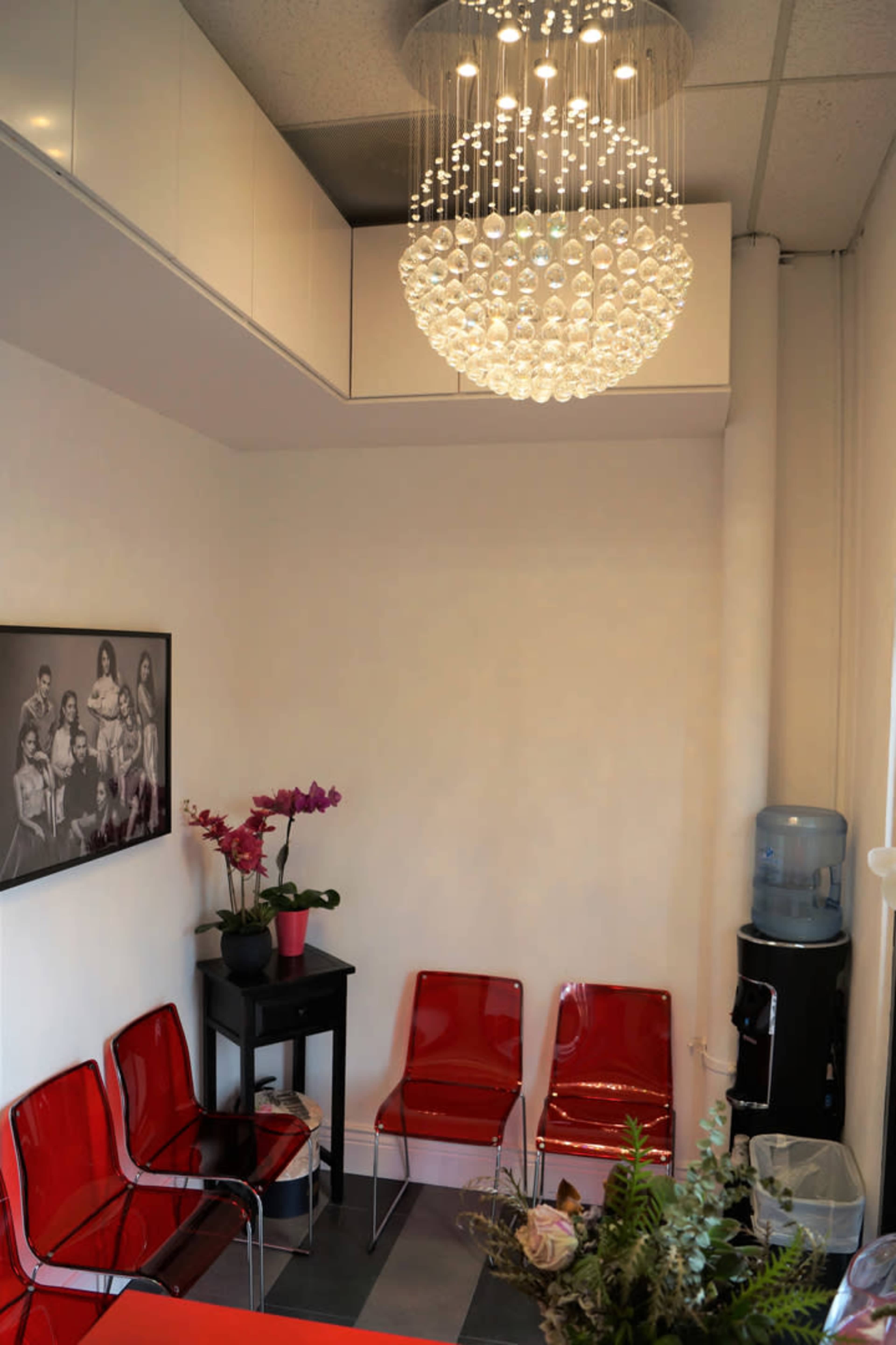 A small waiting area features red transparent chairs, a black side table with an orchid, and a large chandelier above, along with a water cooler in the corner.