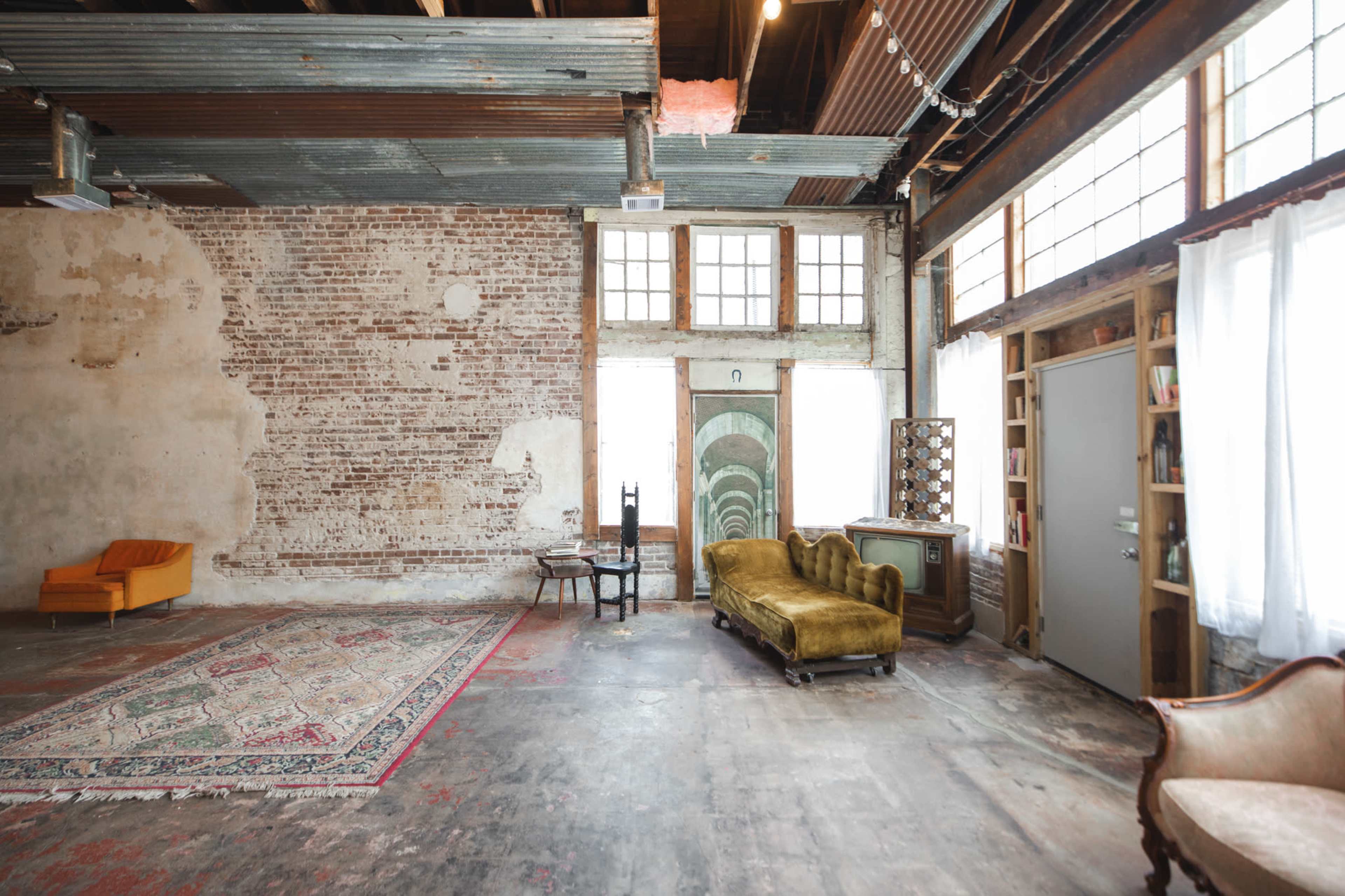 An industrial-style room with exposed brick walls, wooden beams, and various furniture pieces, including a yellow chair and a green couch, arranged around a large area rug.