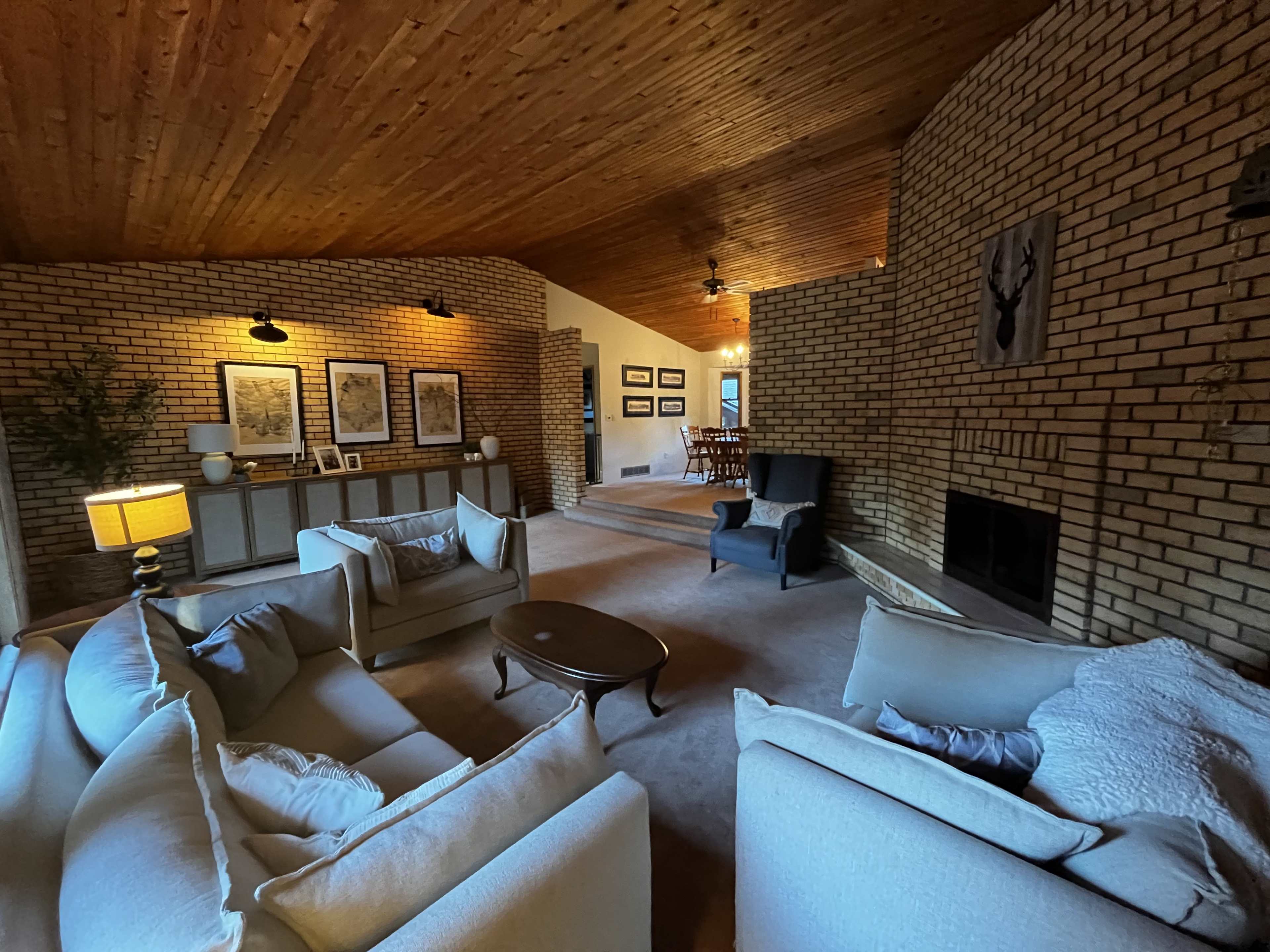The living room features a high wooden ceiling, brick walls, a fireplace, and two sofas facing a coffee table.