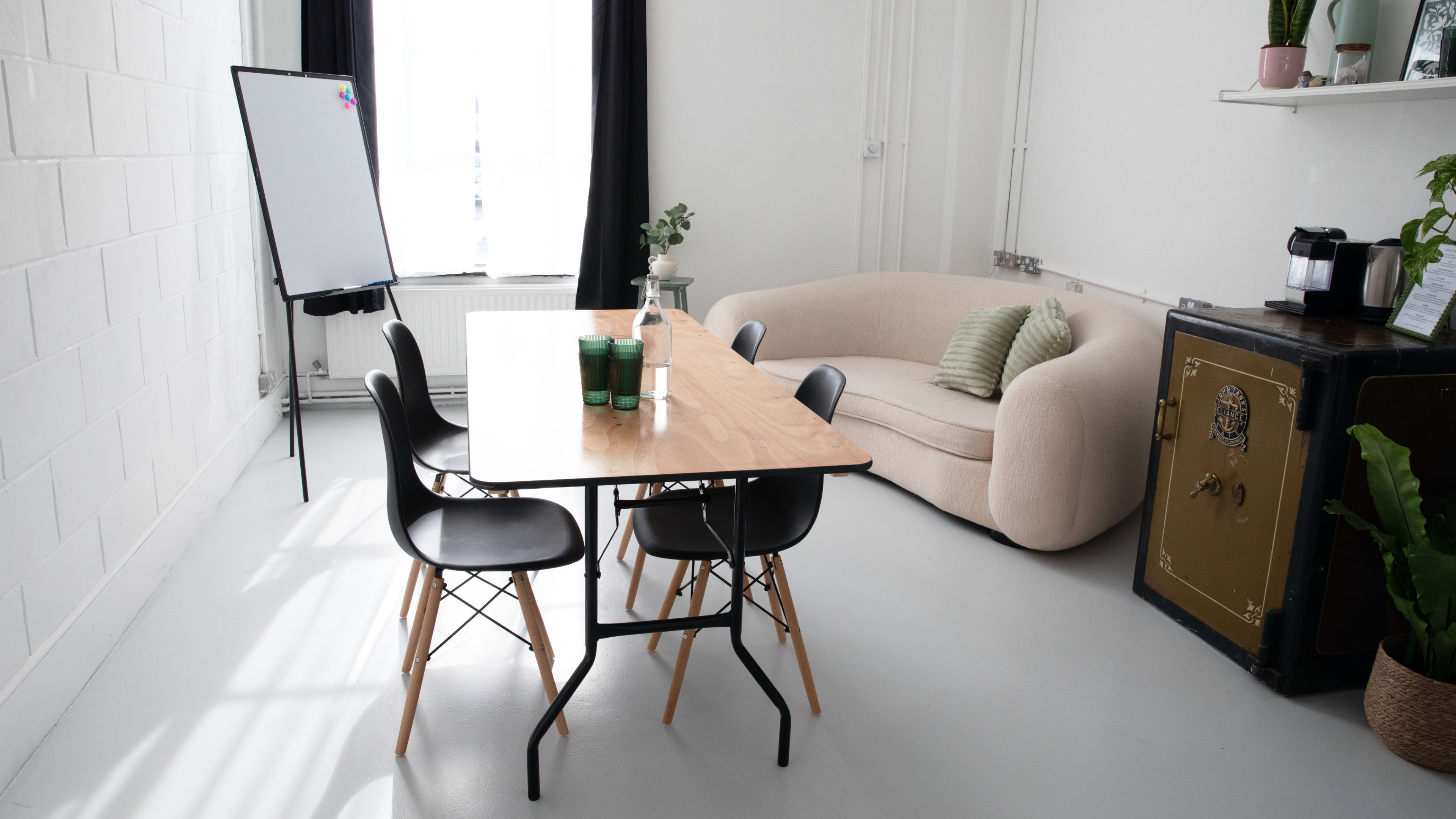 A minimalist meeting room features a wooden table surrounded by black chairs, with a whiteboard and a couch in the background.