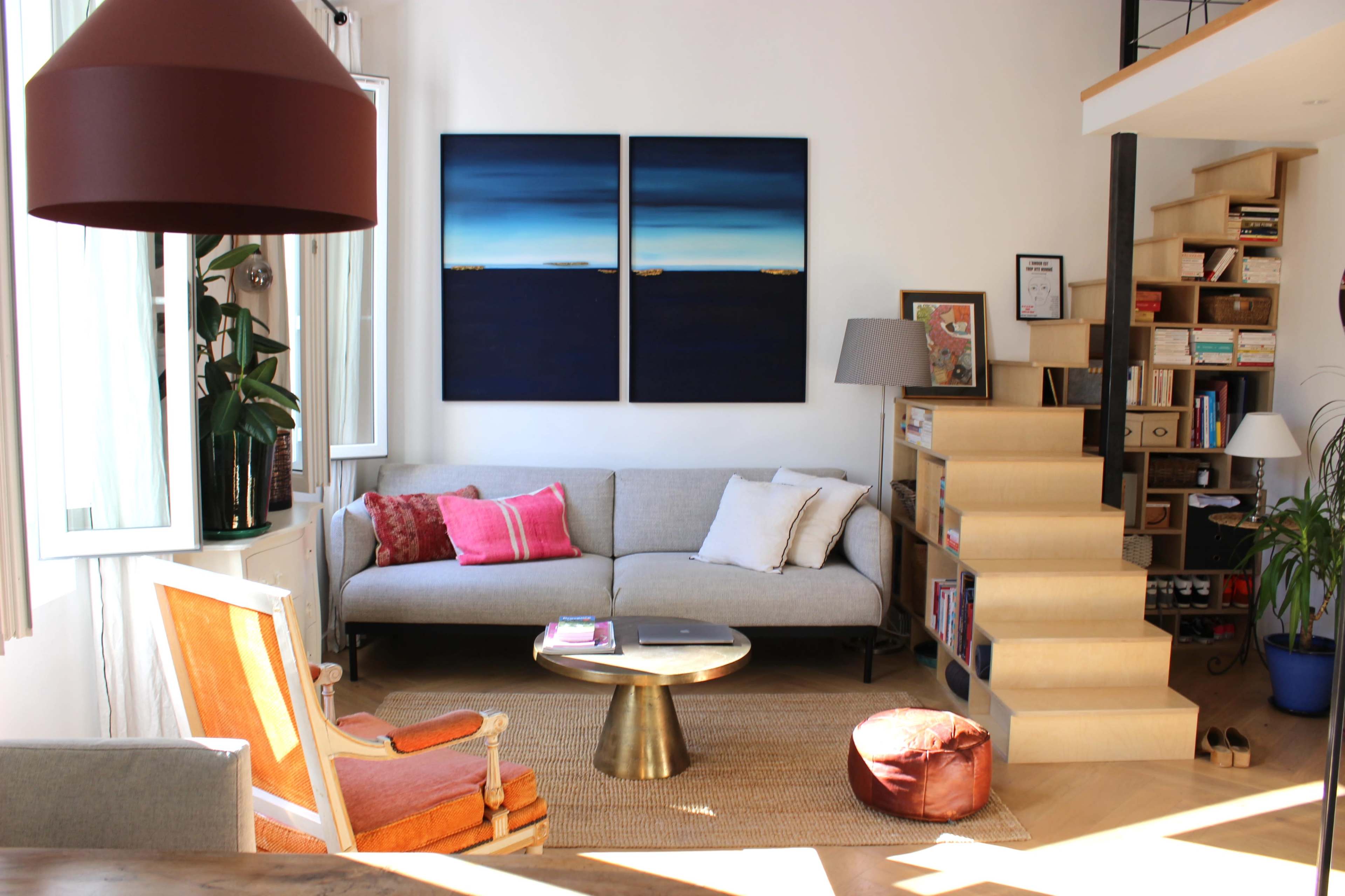 The image shows a bright living room with a gray sofa, colorful cushions, a round gold coffee table, and wooden bookshelves, alongside a staircase leading to an upper level.