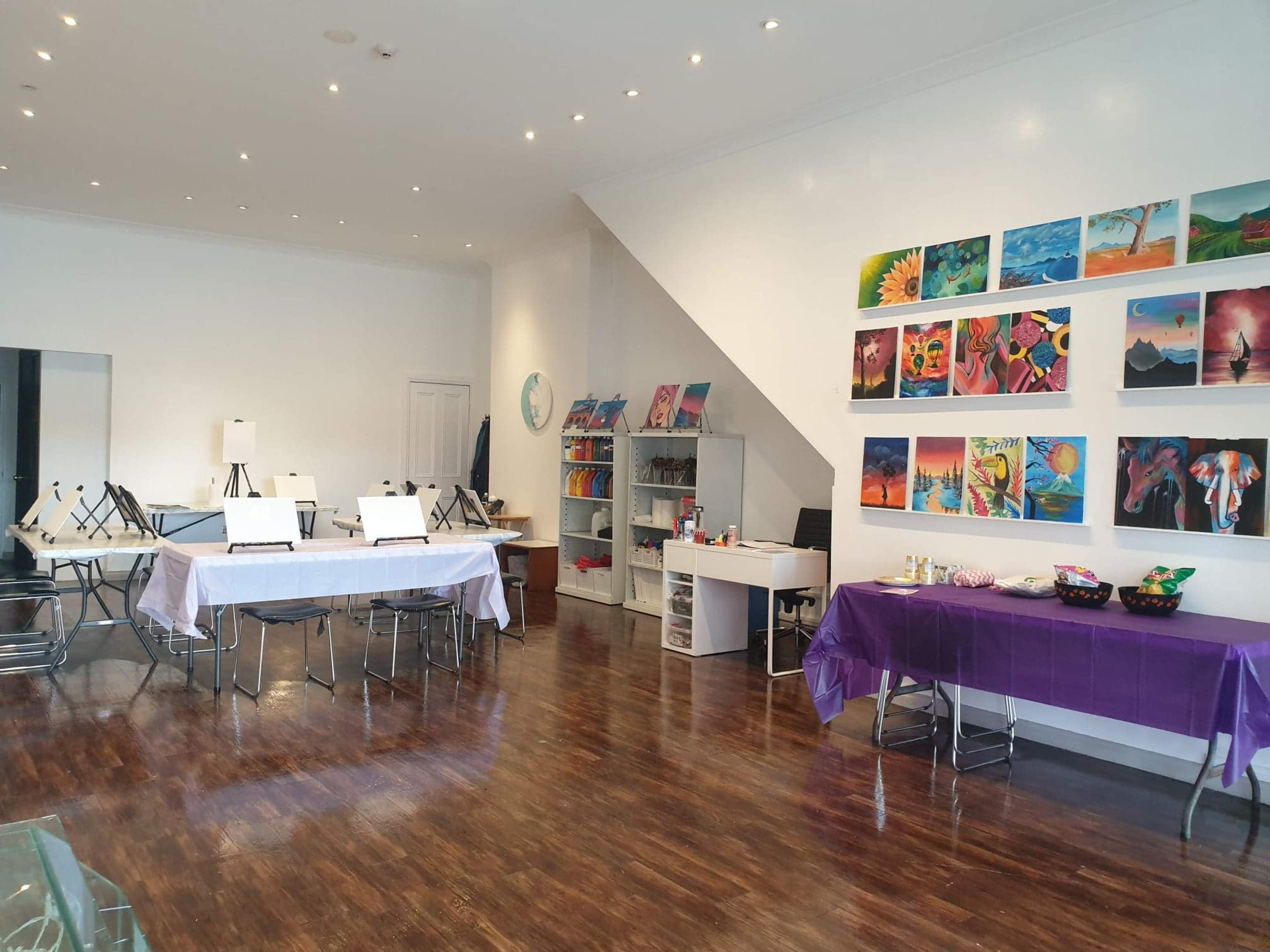 A well-lit art studio features several tables set up with easels, an assortment of completed artwork on the walls, and supplies organized in cabinets.