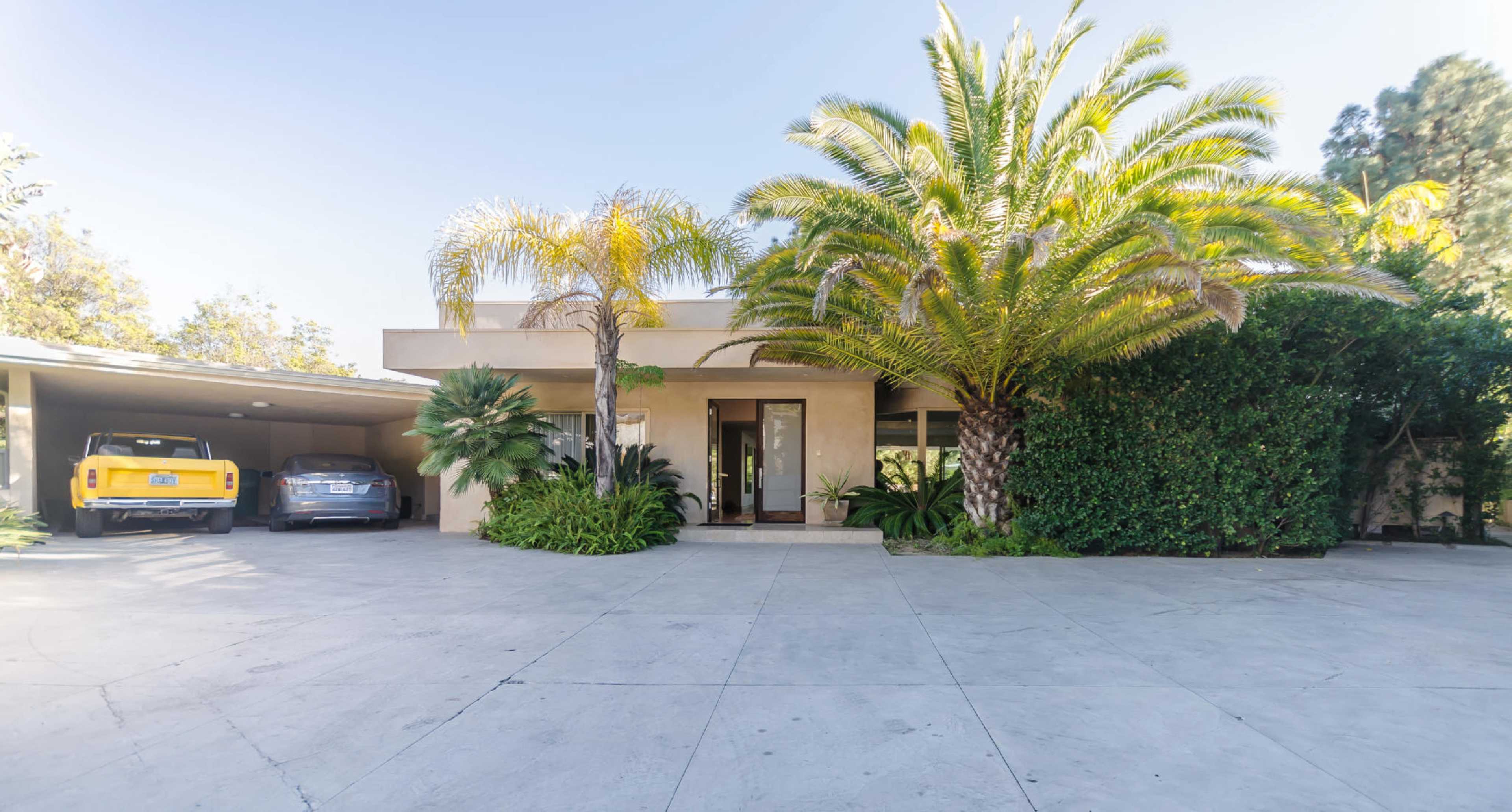 A modern house with a concrete driveway features two parked cars and tropical palm trees in the front yard.